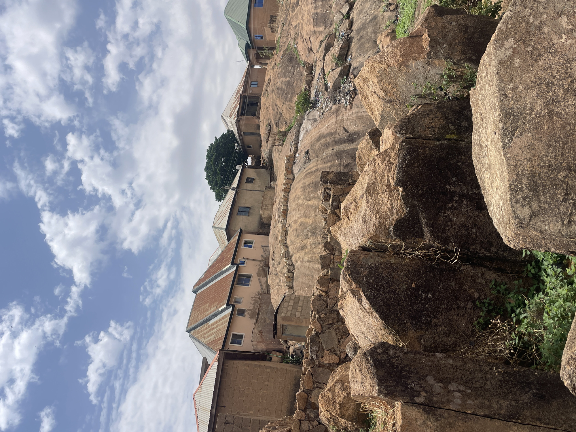 The image shows a hillside community in Jos, Nigeria. Buildings with tin roofs are interspersed among large, dark tan boulders. The architecture is simple, with unpainted, block-like structures. The sky is partly cloudy. A single tree is seen among the buildings. There are visible signs of trash scattered along the hillside.