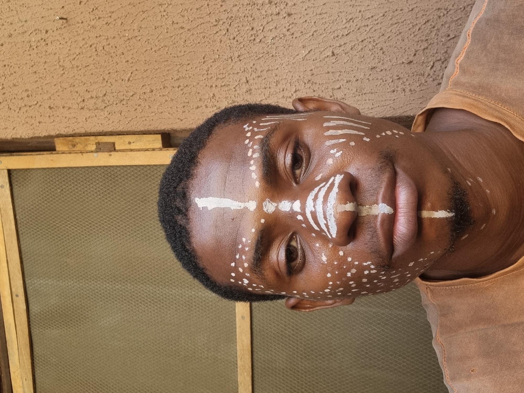 A close-up portrait shows a man with dark skin, adorned with white paint designs on his face. The designs consist of lines on his forehead and nose, and dots on his cheeks and chin. He has short, dark hair and dark eyes. He is wearing a light brown shirt. The background features a beige, textured wall and a window screen with a wooden frame. The location and city are not identifiable in the image.