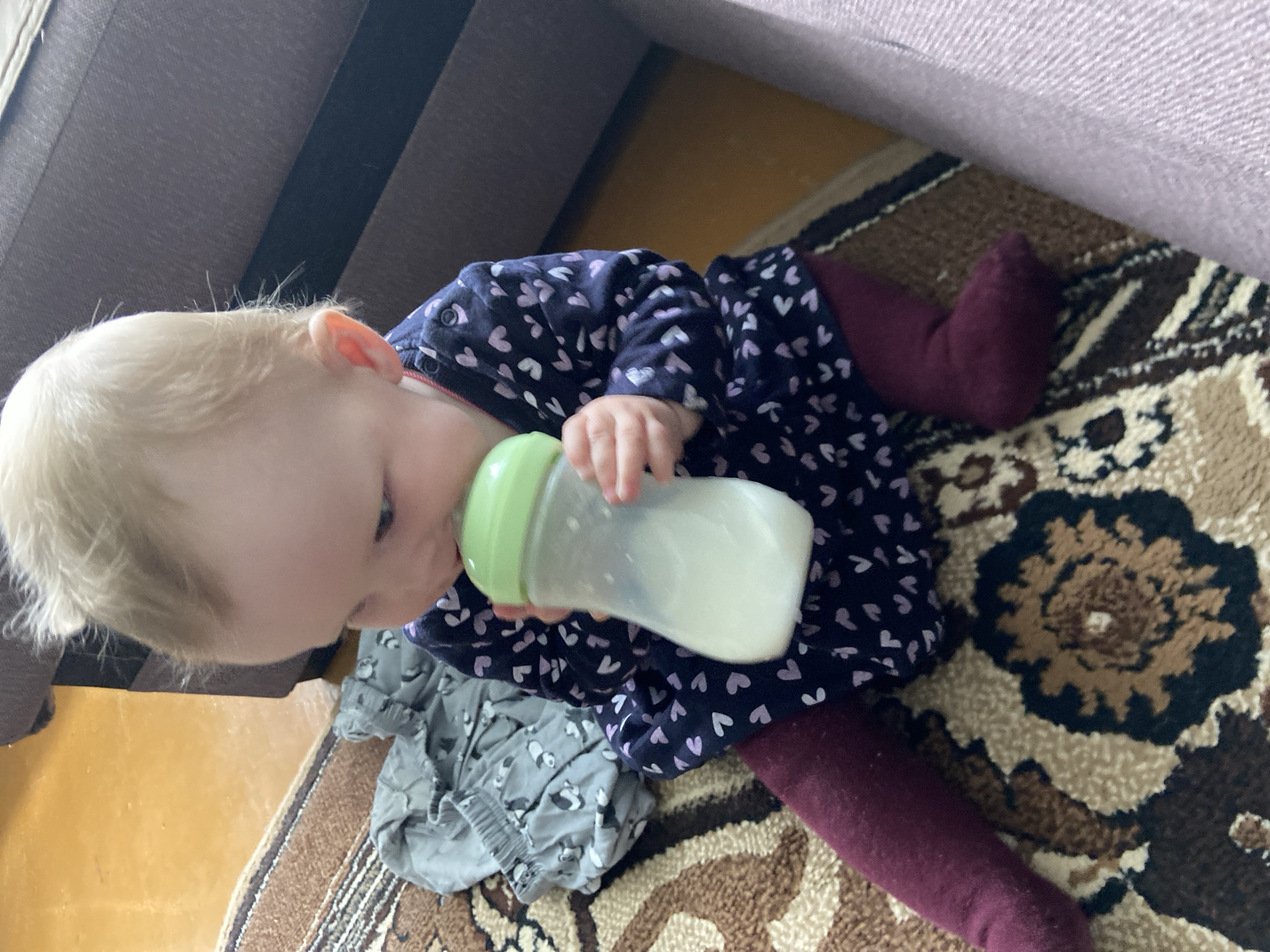 A baby is lying on a patterned rug while drinking from a bottle. The baby is wearing a navy blue dress with white hearts and maroon leggings. There is also a gray patterned garment near the baby. Parts of a couch are visible in the top corners of the photo. It is impossible to determine if this image was taken in Фоки, Russia.