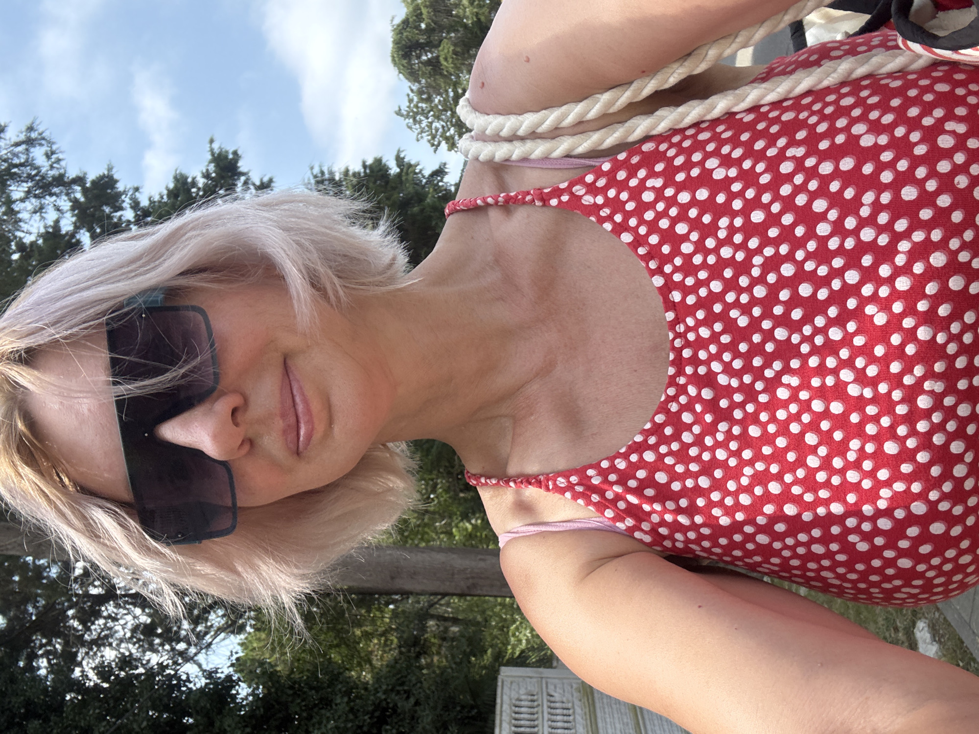A woman with blonde, shoulder-length hair, wearing large dark sunglasses and a red and white polka-dot tank top, is taking a selfie outdoors. She appears to be standing on a paved area, with greenery and trees in the background, under a bright, cloudy sky. A thick rope strap, likely from a bag, is visible over her right shoulder. The scene suggests a casual outdoor setting, possibly in a park or residential area, which could be consistent with the environment found in Sochi, Russia, a city known for its blend of urban and natural landscapes.