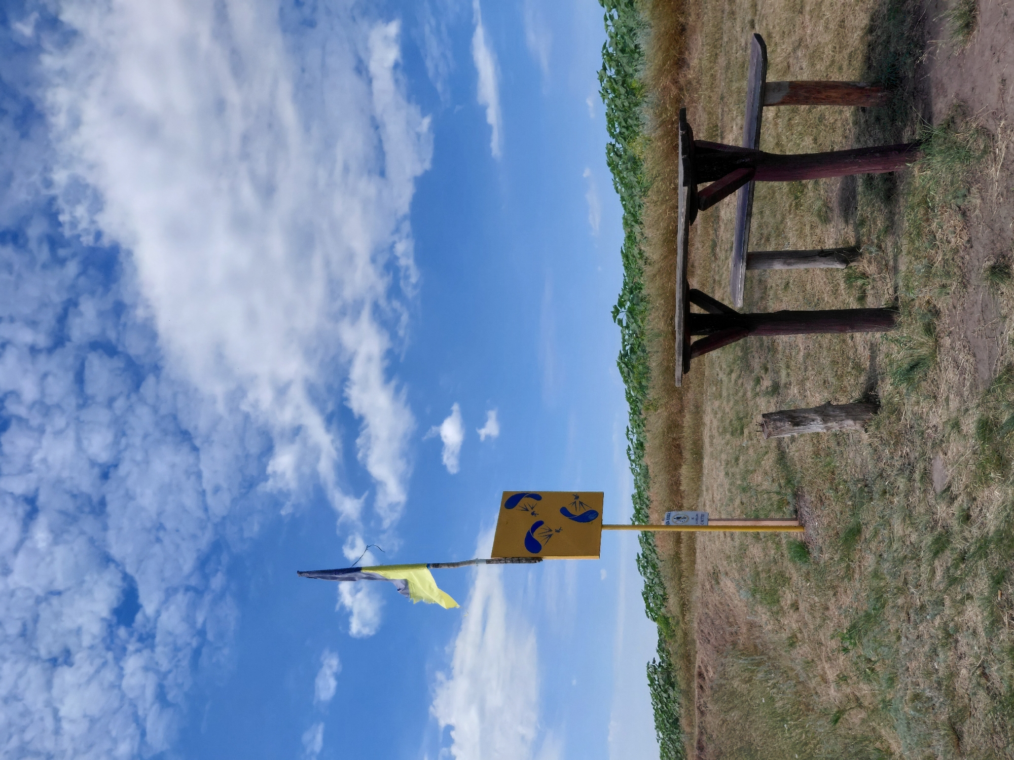 A vertical, medium shot captures an outdoor scene with a blue sky dotted with white clouds. In the foreground, a rustic wooden picnic table and bench are set on dry, patchy grass. Behind the table, a yellow signpost with a Ukrainian flag, tattered and waving, stands near a field of green plants. The signpost features a yellow square with three blue icons of paragliders. A small white sign with text is attached to the lower part of the post. The terrain slopes gently upwards, with dry vegetation and sparse grass. The background reveals a vast field of green plants, likely sunflowers, under a bright sky. The overall impression is of a rural or recreational area.