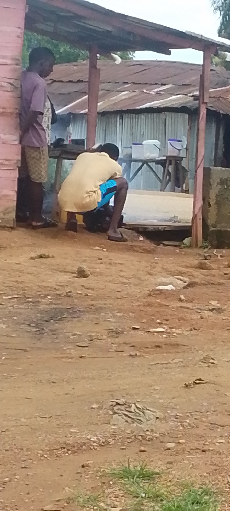 The image shows an outdoor scene in Baissa, Nigeria. In the foreground, there is a dirt ground with scattered stones and some sparse vegetation. A person wearing a yellow t-shirt and blue shorts is crouching down, appearing to be working on something on the ground. To the left, another person, seen from the waist down, is standing and leaning against a pink structure. Behind these individuals, there is a rustic shelter with a corrugated metal roof supported by wooden posts. On a table behind the crouching person, several containers, possibly buckets, are visible. The overall environment suggests a market or a residential area with informal structures.