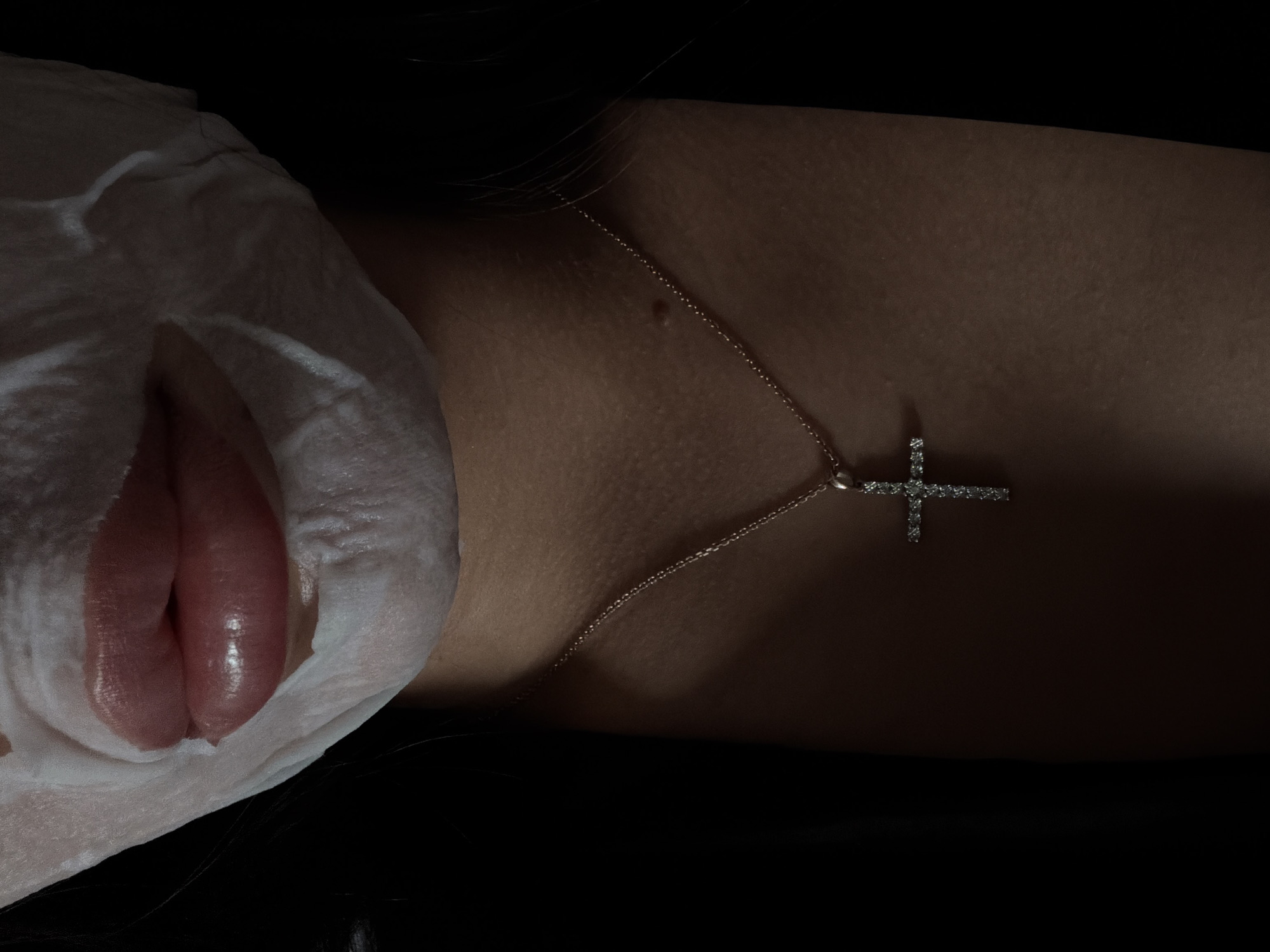 The image shows a person lying down with a facial sheet mask applied. The mask covers most of their face, except for their mouth. They are wearing a delicate chain necklace with a cross pendant. The background is black and indistinct. There is no information in the image to suggest the location of the image is Krasnoyarsk, Russia.