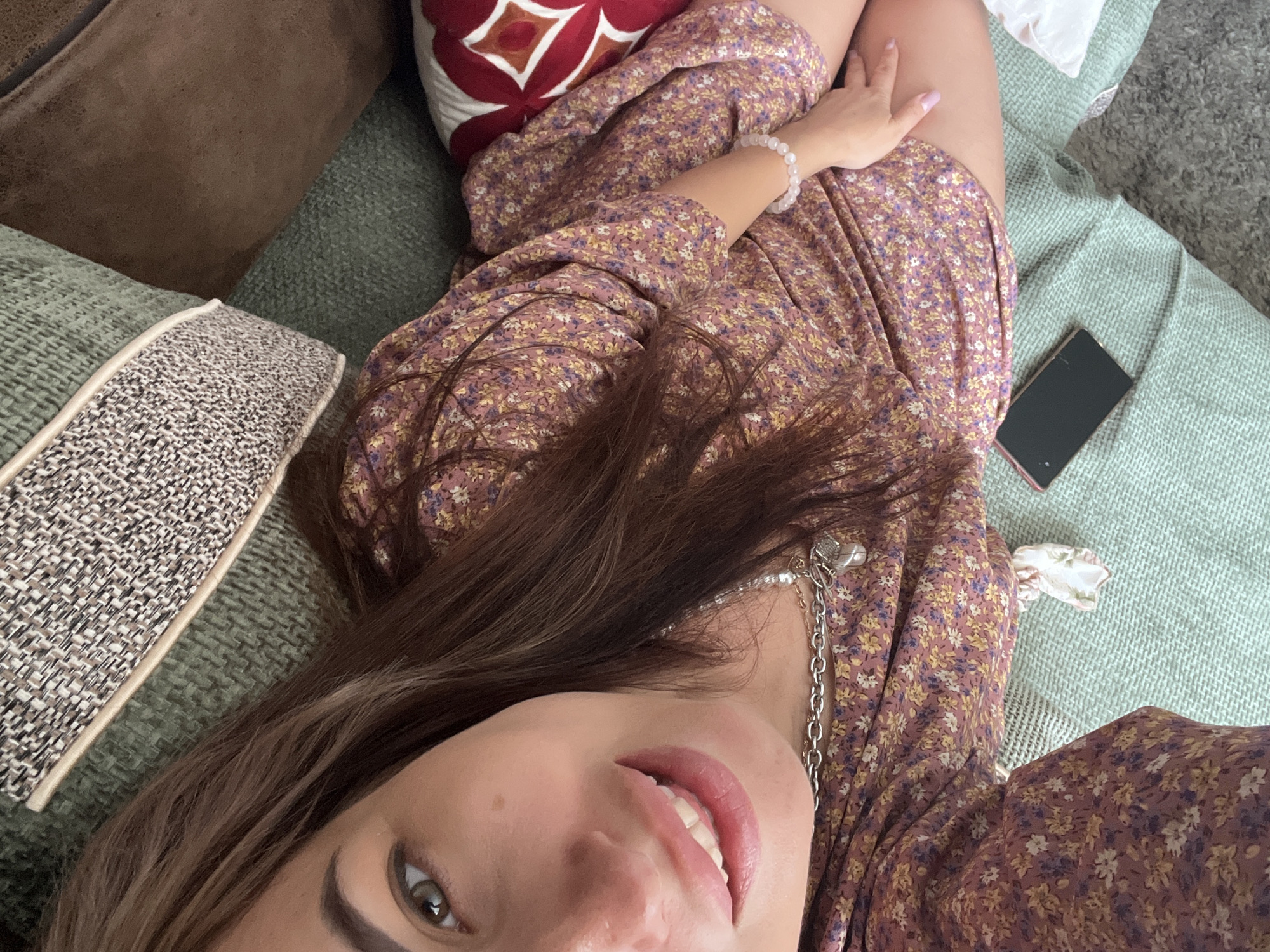 A young Caucasian female with long, dark brown hair and light green eyes is lying on a couch. She is wearing a short, floral print dress and a silver chain necklace. Her left hand is resting on her thigh, and she is smiling. A cell phone is visible on the couch next to her. The couch appears to be in an indoor setting, possibly a living room, with textured pillows and throws in various shades of green and brown.