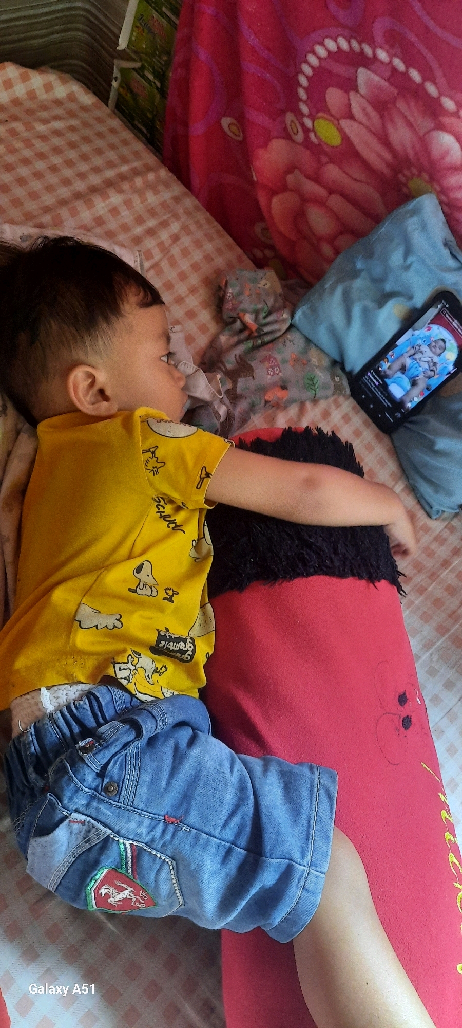 The image shows a young child sleeping on a bed. The child is wearing a yellow t-shirt with cartoon characters and blue denim shorts. The child is lying on their side, facing away from the camera, with their arm resting on a red pillow. A smartphone is placed next to the child, displaying a video of a baby. The bed is covered with a pink and white checkered blanket and a brightly colored pink blanket with a floral pattern. The setting appears to be a bedroom in a home, possibly in Ploso, Indonesia, given the location context. The lighting suggests it is daytime. The overall mood is peaceful and quiet, with the child seemingly having fallen asleep while watching a video. There is a visible watermark "Galaxy A51" in the bottom left corner of the image.