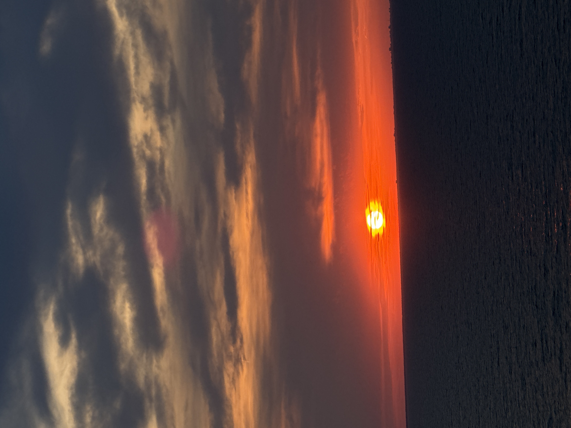 The image depicts a sunset over a body of water, possibly the sea. The sky is filled with clouds in various shades of orange, red, and dark gray, with the sun appearing as a bright orange orb on the horizon. The water is dark and appears calm, with subtle ripples. A faint line of buildings can be seen on the distant horizon, suggesting a coastal city. The scene is natural and serene, capturing the dramatic colors of a sunset. The location is listed as Unknown, Italy, but the image itself does not provide specific geographical identifiers.