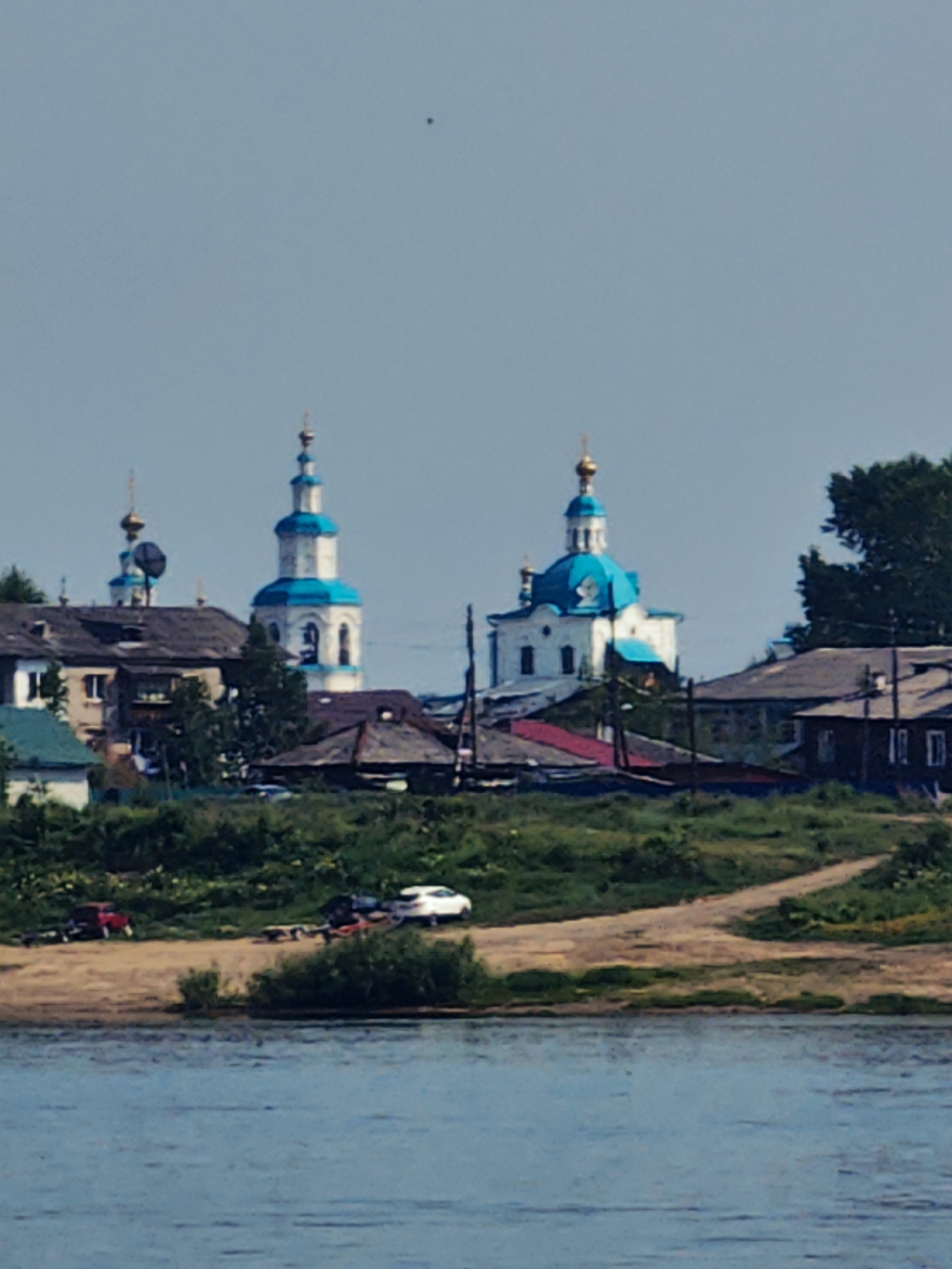 A distant, slightly out-of-focus view of the city of Yeniseysk, Russia, showcases a waterfront scene. The foreground features a body of water, likely the Yenisei River, with ripples suggesting a gentle current. Along the riverbank, a sandy shore is visible, with a path winding through green vegetation and some bushes.
Further inland, a cluster of buildings forms the cityscape. Dominating the view are two distinctive churches with white walls and vibrant blue domes, topped with golden crosses. One church is taller and more prominent, featuring a bell tower. Adjacent to the churches are residential buildings with darker roofs and visible windows. Some of these appear to be older, wooden structures, while others are more modern.
On the sandy bank, two vehicles are parked. A red car is on the left, seemingly attached to a trailer. To its right, a white car is also parked, also with a trailer. The overall impression is one of a peaceful, somewhat rural urban setting. The sky is a pale blue, with a single small dark speck visible high up, possibly a bird or an insect.