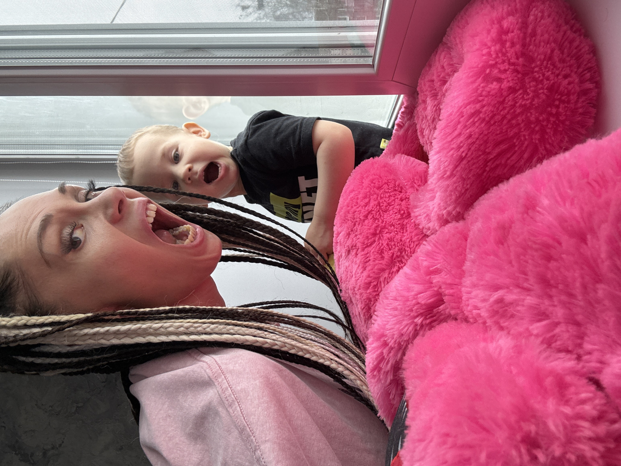 A low-angle shot depicts a woman and a young child looking down into the camera. The woman is Caucasian, with long braids, wearing a pink top, and she has her mouth open wide. The child, also Caucasian, is wearing a black shirt with yellow lettering. Both subjects appear to be in a state of excited expression. Behind the child, a large pink, fluffy object is partially visible. In the upper background is a window with blinds. The location of the photo may be Rusanovo, Russia.