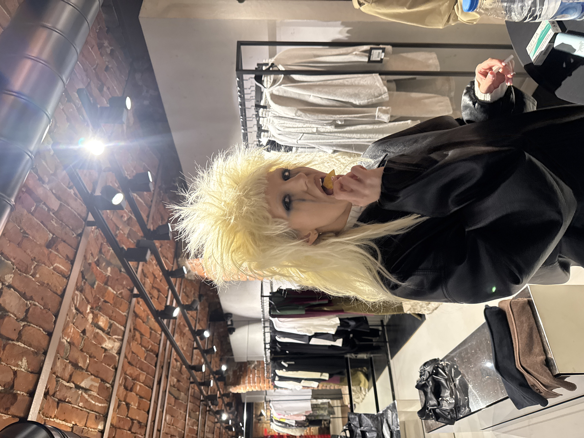 The image depicts an individual standing within a retail establishment located in Sofia, Bulgaria. The central subject, a person with voluminous, spiky, bleached blonde hair, faces the viewer directly. They wear heavy black eyeliner, with prominent black streaks extending downwards from the outer corner of the left eye, resembling smudged mascara or tears. The individual is wearing a black, possibly leather, jacket or coat over a visible white garment at the wrist. Their right hand holds a small, irregular yellow-orange object, which they are in the process of biting or placing into their open mouth.
In the foreground, to the individual's right, a black countertop holds a clear plastic water bottle, a small white and green box labeled "DELIA," and a light blue rectangular object. To the left in the mid-ground, a reflective, possibly metallic, display table features a black leather handbag with metallic accents, along with two folded items, one black and one brown, possibly gloves or scarves.
The background reveals the interior of the store, characterized by an exposed arched brick ceiling with light-colored mortar. Modern black track lighting fixtures with multiple spotlights are affixed to metallic beams running across the ceiling. A large, metallic ventilation duct is also visible near the ceiling. Numerous clothing racks display garments; those on the left feature a variety of colors and styles, while racks on the right primarily hold light-colored (grey, beige, white) casual clothing items such as sweatshirts or long-sleeved shirts, some with visible white tags. Sections of exposed brick walls are also discernible further into the store.