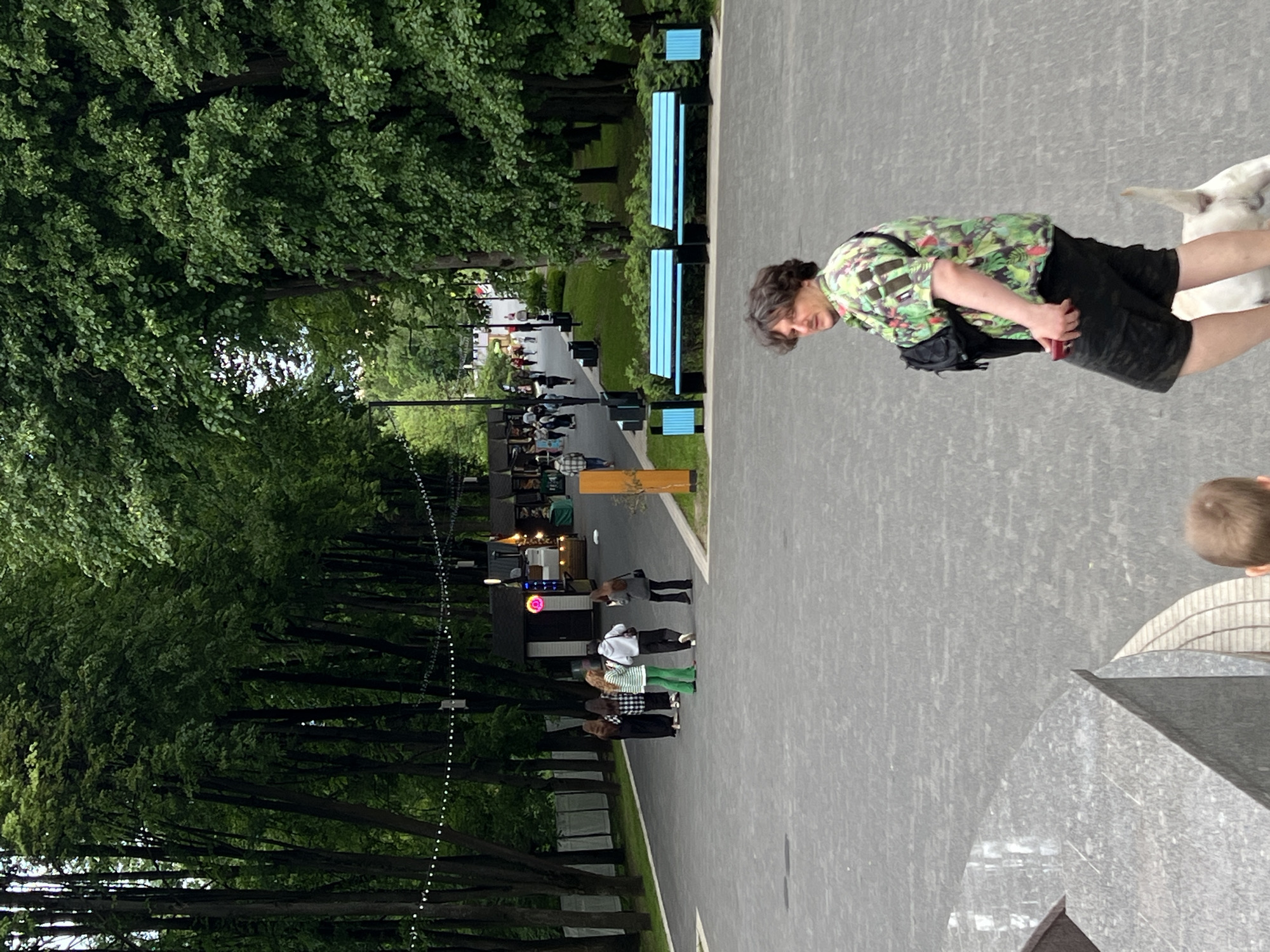 A photograph captures a well-lit outdoor scene. A person in a green patterned shirt and dark shorts stands on a paved area. A white dog is present. In the background, a pathway is lined with trees on the left. People are walking along the pathway. A row of blue benches sits to the right of the path. The location appears to be a public space in Mytishchi, Russia.