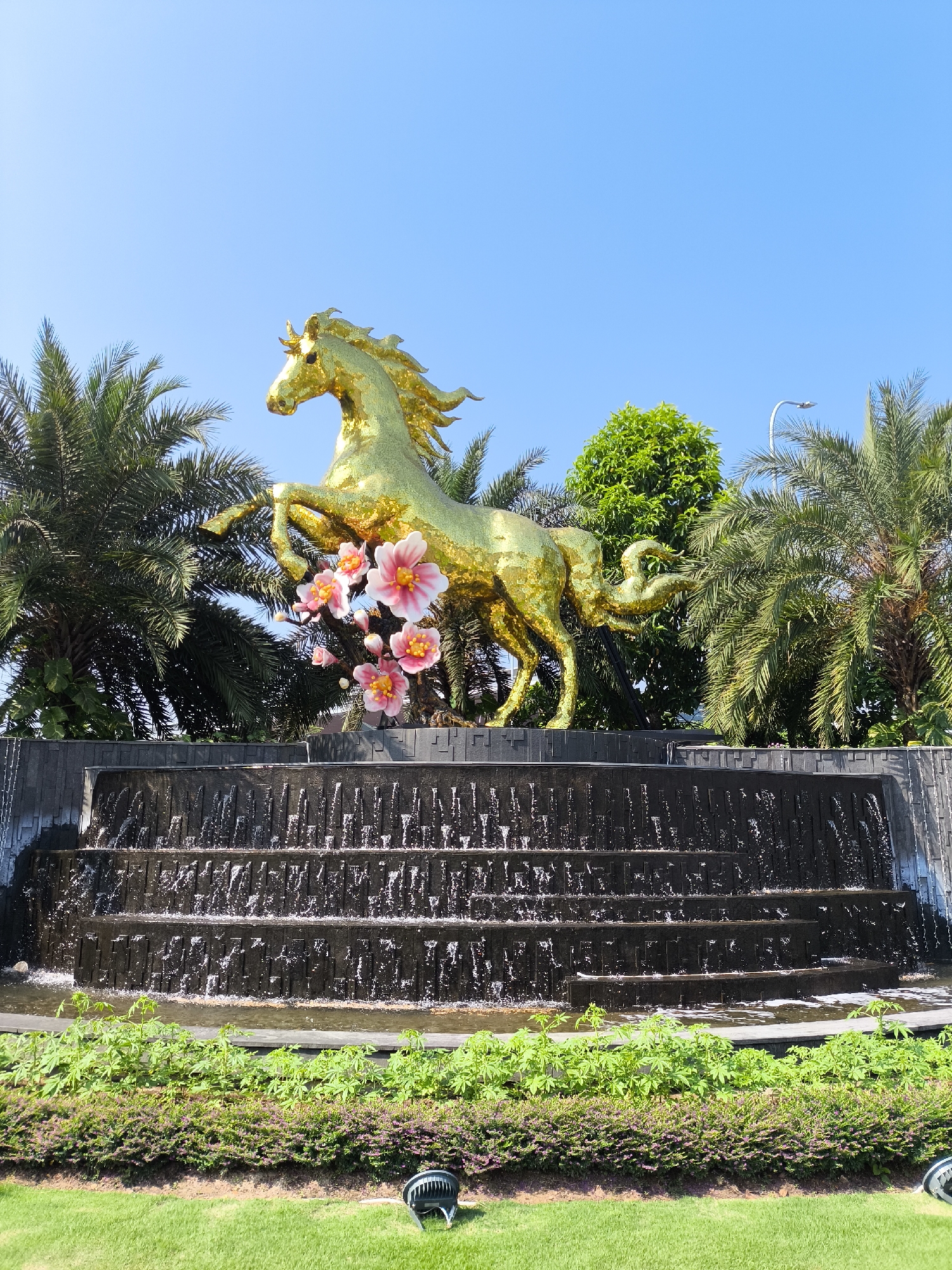 An outdoor scene in Đà Nẵng, Vietnam, features a prominent golden horse statue positioned atop a dark, multi-tiered waterfall structure. The statue, depicted in a rearing pose facing left, is adorned with several clusters of artificial pink and yellow flowers near its raised front left leg. Water cascades down the various levels of the dark, rectangular fountain base. Surrounding the statue and fountain are numerous tall palm trees and dense green foliage under a clear, light blue sky. In the foreground, a manicured green lawn is visible, separated from the fountain by a row of low green shrubs and smaller leafy plants. A black, circular floodlight is present on the grass in the lower right quadrant. No individuals are visible. This landmark is located at the intersection of Võ Nguyên Giáp and Võ Văn Kiệt streets in Đà Nẵng.