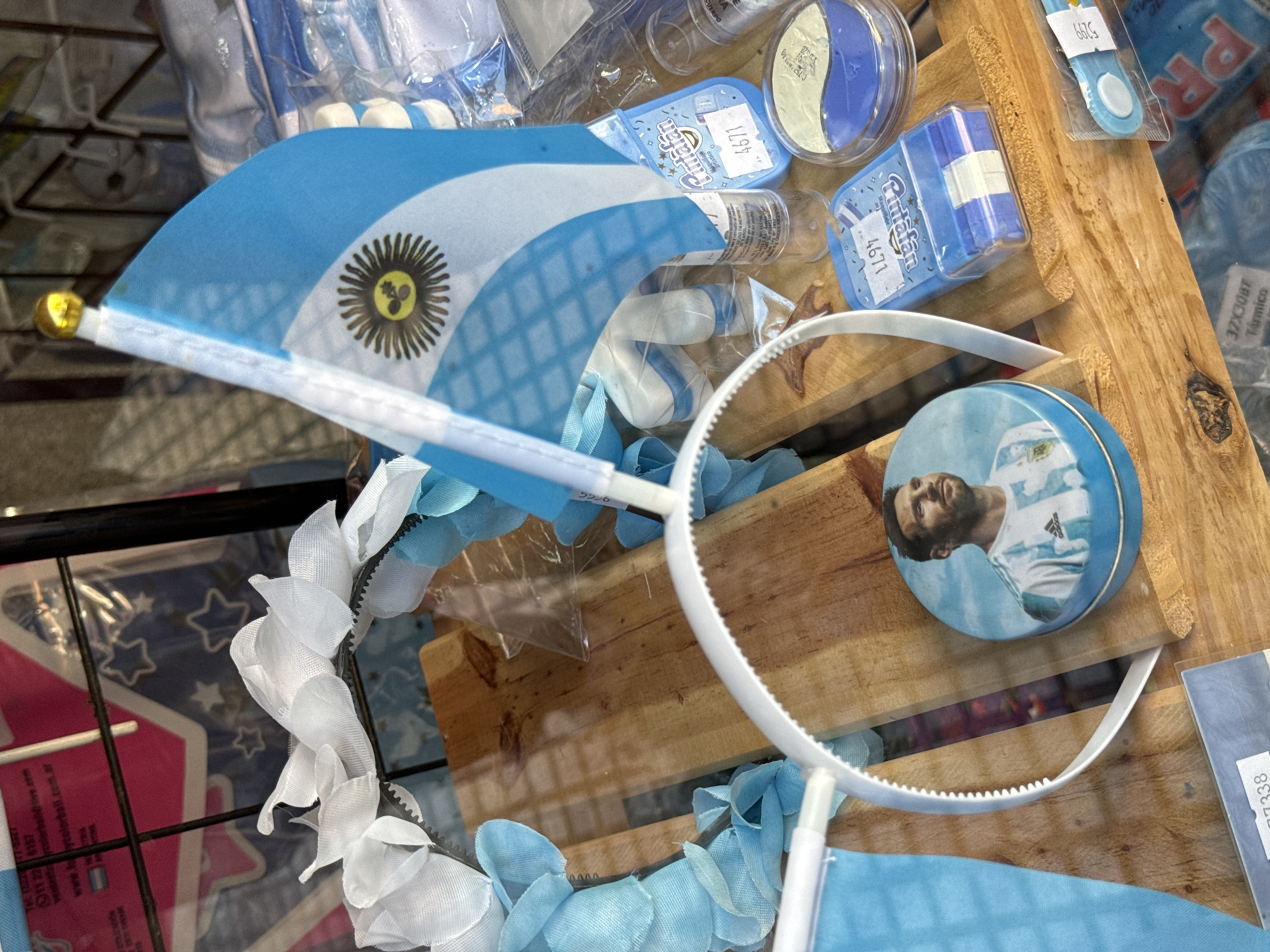 This eye-level, close-up shot displays a collection of Argentina-themed merchandise. The items are displayed on a wooden shelf within a wire rack, likely in a shop or market in Buenos Aires, Argentina.
An Argentina flag is prominent, featuring the iconic light blue and white stripes and the sun emblem. Near the flag is a white headband adorned with white floral accents, and another headband, decorated with blue fabric, with a tin featuring the image of soccer player Lionel Messi on the wood.
Various containers of "Rutafan" brand face paint, with blue and white colors mirroring the Argentine flag, sit among the other items. The containers have labels with "4671" and other codes printed on them. The labels of the face paint mention "Maquillaje Artistico," which translates to artistic makeup. One label bears the information "11 22 6157" and a website "www.colorball.com.ar," possibly indicating contact details for the brand. Also present is a label reading "37X30BT Termico".