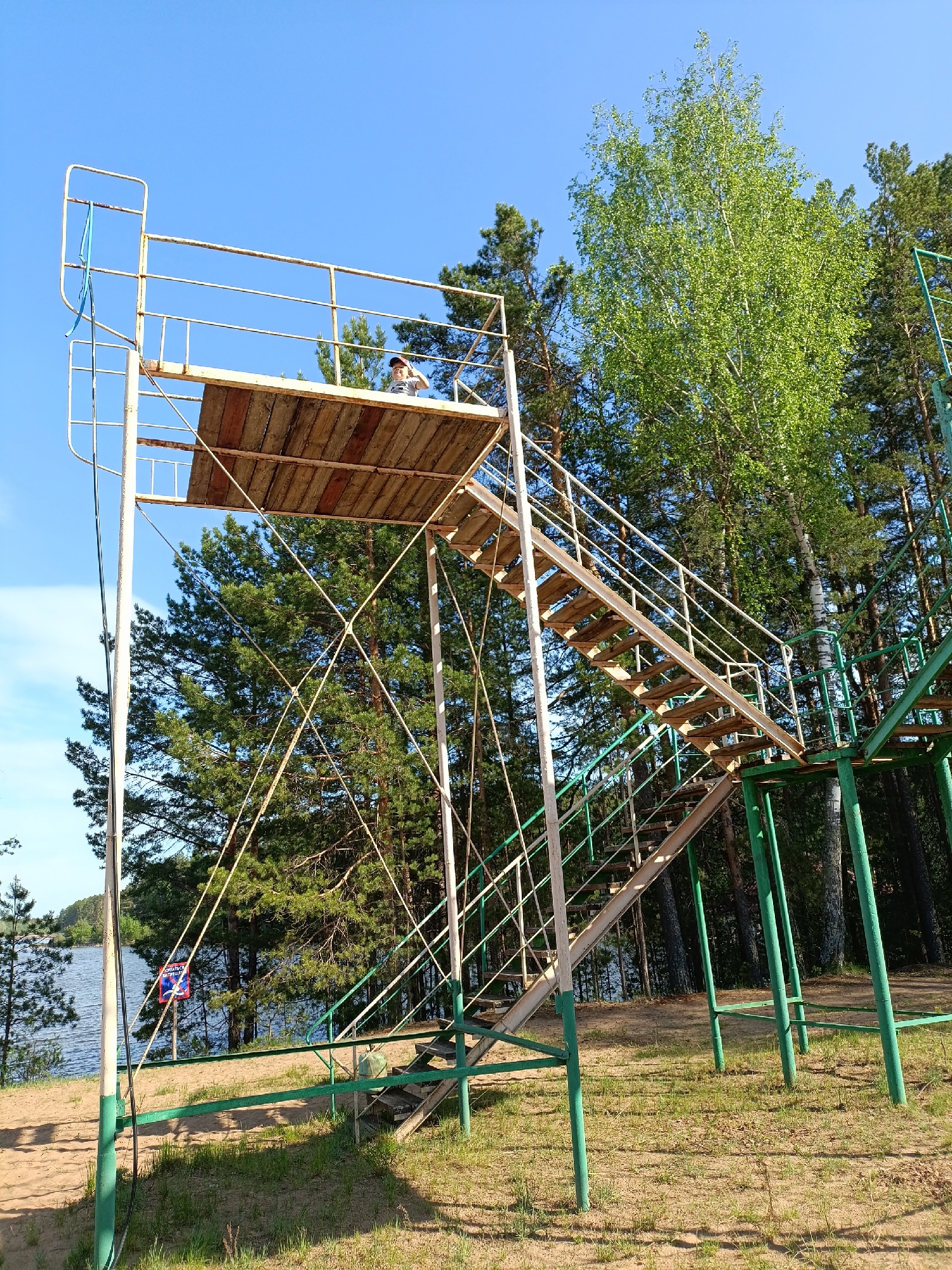 A ground-level outdoor shot shows a high diving platform on the shore of a body of water, assumed to be the Kama Reservoir in Chaykovsky, Russia. The platform is composed of a wooden platform at the top, accessed by two flights of metal stairs with railings. The supporting structure is a mix of metal beams and supports, some painted green. The ground is a mix of sand and grass. In the background are trees, including evergreens and a leafy tree. A small figure can be seen on the top platform. A blue life preserver hangs near the base of the platform. The sky is clear blue.
