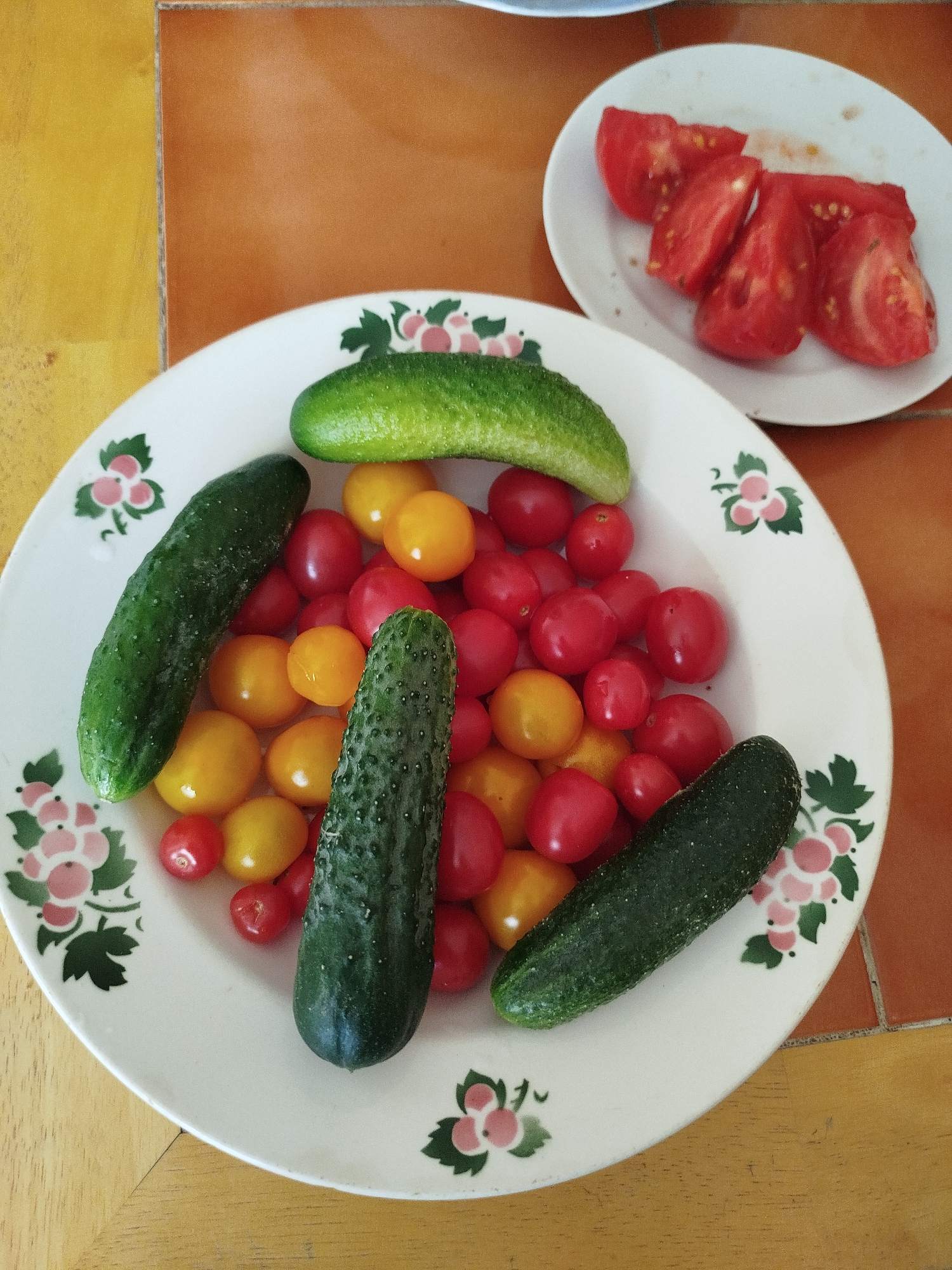 A close-up, high-angle shot shows a plate of cherry tomatoes and cucumbers on a table. The plate has a floral pattern with pink and green accents. There are at least three cucumbers and numerous red and yellow cherry tomatoes. To the upper right, on a separate white plate, are several slices of chopped red tomato. The table appears to be made of wood, with a tile inlay in the upper portion of the image. The setting does not provide any information about the city of Pskov, Russia, or any people present.