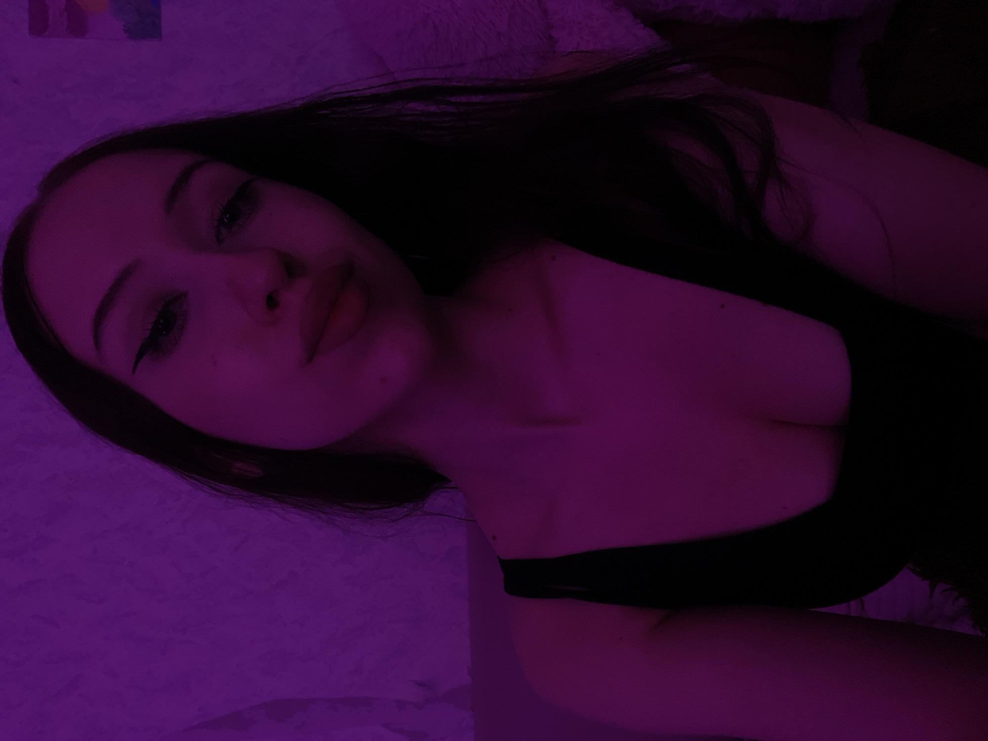 The image is a selfie taken in a dimly lit room, illuminated by purple lighting. The subject, a young woman with dark hair and dramatic winged eyeliner, is looking directly at the camera. She is wearing a black tank top that reveals her décolletage. The background is a textured purple wall, with what appears to be a small piece of art on the upper right side. The overall tone of the image is intimate and moody. There is no indication of the city of Votkinsk, Russia, or any specific location within it.