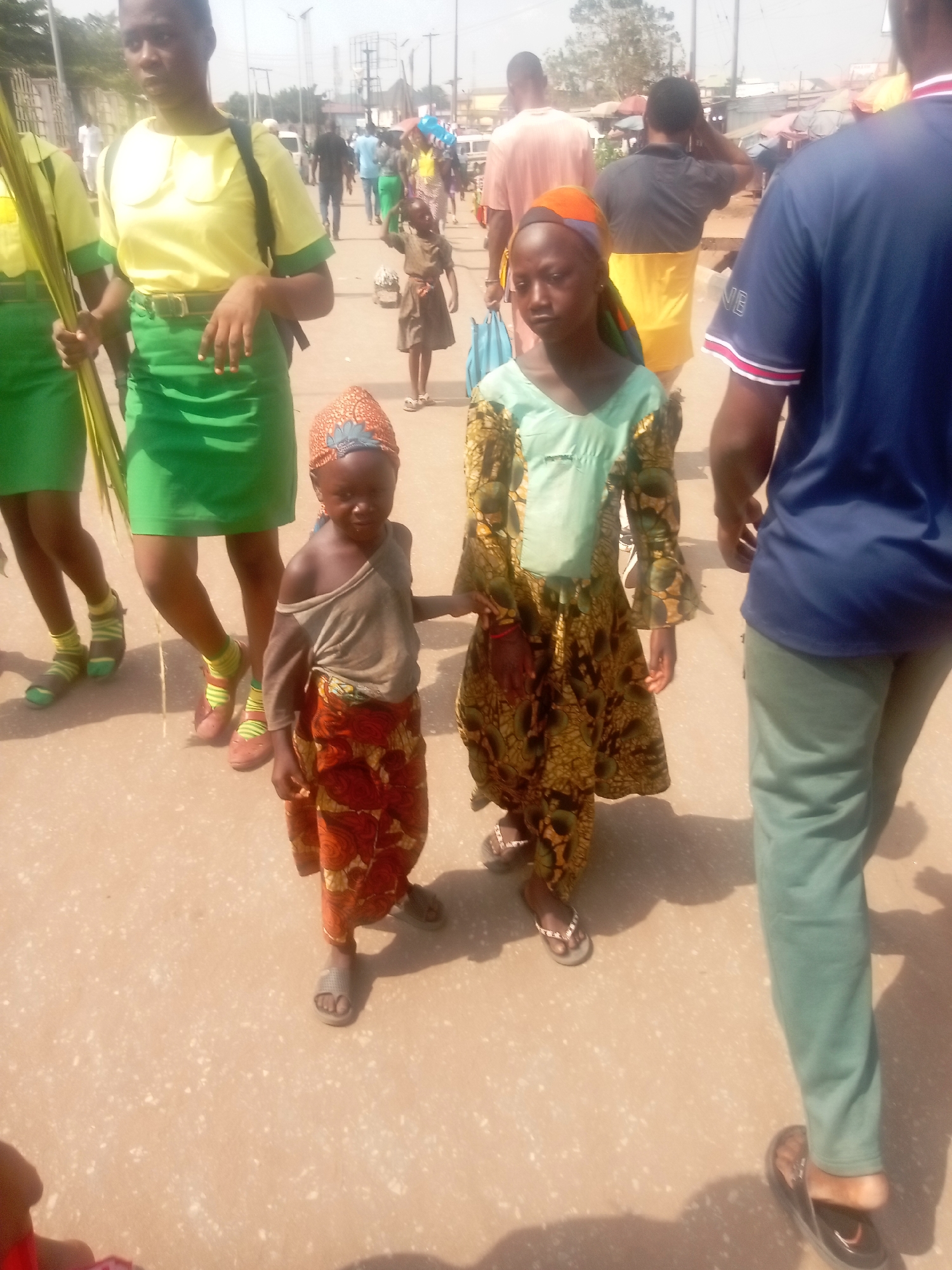 The image depicts a daytime street scene in Onitsha, Nigeria, with multiple individuals walking on a light-colored paved surface under direct sunlight.
In the foreground, from left to right:
A young female, partially visible, wears a yellow short-sleeved shirt with a green collar and sleeve trim, a green skirt, and a black backpack. She holds several long, green palm fronds vertically in her left hand. Her lower legs show striped green and light brown socks with brown/red sandals.
To her right, a small child, facing forward, wears a reddish-orange patterned headscarf, a light brown/grey long-sleeved shirt with one shoulder exposed, and a red/orange patterned skirt. This child wears grey slip-on sandals.
Adjacent to the first child, another child, also facing forward, wears an orange and blue patterned headscarf, a light teal v-neck top, and a long gold/green patterned dress with bell sleeves. This child wears light-colored flip-flops. The two children appear to be standing or walking near each other.
On the far right, the back of a male figure is visible, wearing a navy blue short-sleeved shirt with white and red striped trim on the sleeves, and light green trousers. He wears dark flip-flops.
In the mid-ground and background, numerous other individuals are walking or standing. These include a person in a light pink shirt, a person in a black shirt with a yellow lower half, and a smaller child in a brown dress. The background shows blurred market-like stalls with colored umbrellas, utility poles, and distant buildings, indicating an urban environment. The sky is bright and hazy. Shadows are cast on the ground from the right side of the frame, suggesting the sun's position.