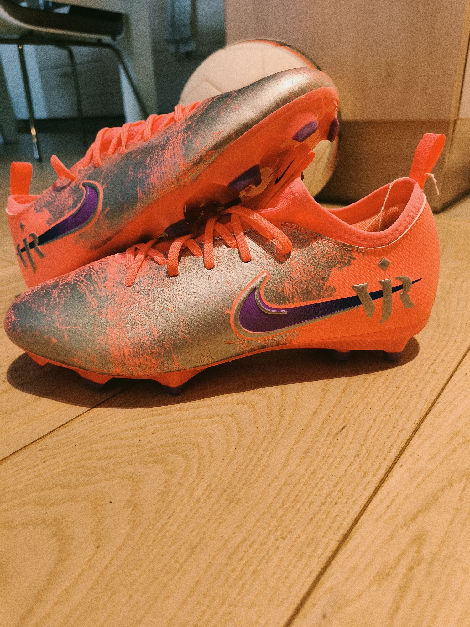 A pair of vibrant coral-colored Nike Mercurial soccer cleats sits atop a light brown wooden surface. The cleats feature a silver-colored overlay with a distressed or cracked pattern in coral. A purple Nike swoosh is visible on the side of each cleat, and stylized initials "VR" are printed near the heel. The cleats have coral-colored laces and a pull tab at the back. Behind the cleats, a partial soccer ball is visible, along with a portion of a light-colored cardboard box. The background also includes glimpses of a chair. The image lacks discernible characteristics that would confirm it was captured in Sofia, Bulgaria.