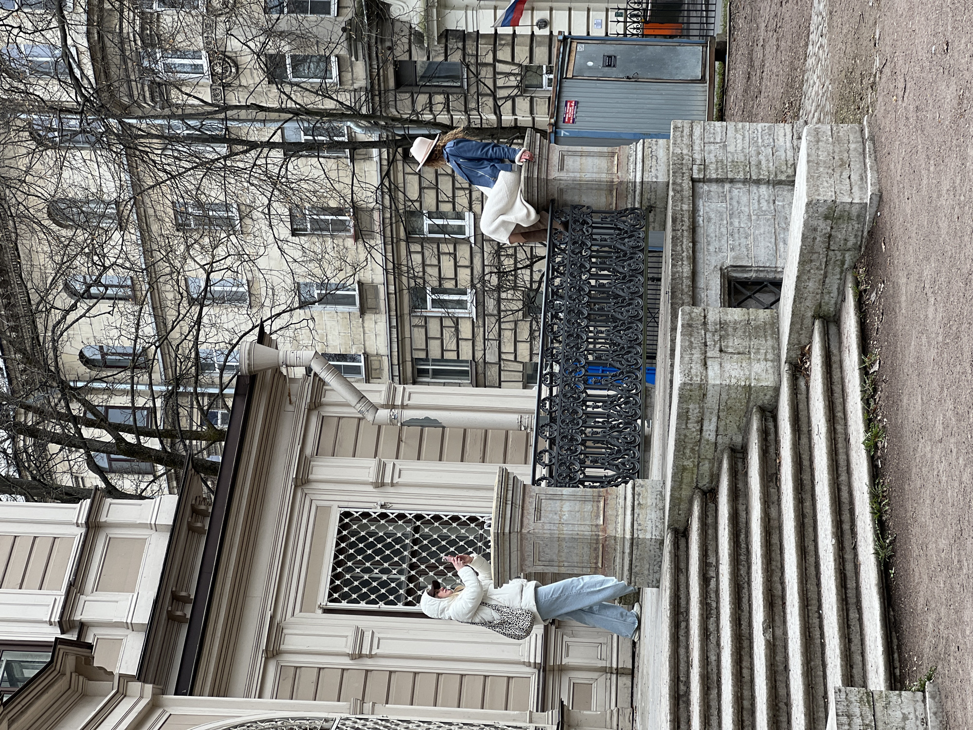 The image shows two people near a building in Saint Petersburg, Russia. One person, wearing a white hooded jacket, light blue jeans, and a shoulder bag with a leopard print pattern, is standing near the corner of the building and appears to be taking a photograph with their phone. The other person, wearing a blue jacket, white pants, brown boots, and a hat, is sitting on a balcony railing. The building features a facade with decorative moldings and a window with a lattice design. There are leafless trees visible in the background. The Russian flag is visible on a building.