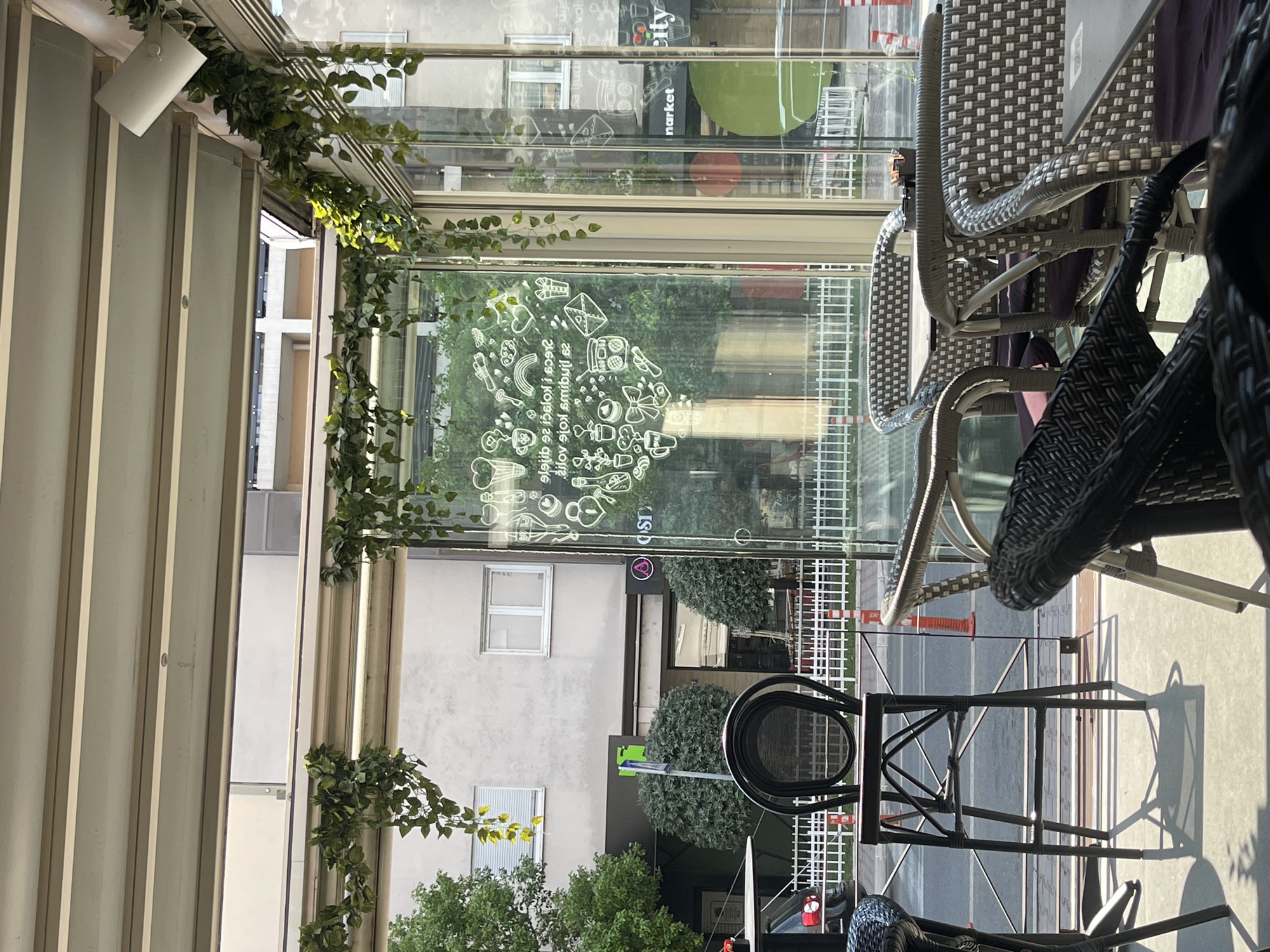 The image depicts the interior of a cafe in Podgorica, Montenegro, looking outwards through a glass-enclosed patio area. The cafe is decorated with hanging green artificial vines along the ceiling beams. Inside, there are several tables and chairs. A tall, black bar stool with a circular seat is positioned prominently.
Through the glass, a street scene is visible. Buildings with light-colored facades and windows are in the background. A white picket fence runs along the edge of the patio. Orange traffic cones are present on the sidewalk. To the right, a sign for "Market City" is visible on a building. The overall impression is that of an outdoor seating area of a cafe on a sunny day, with everyday city life in the background. There are no people clearly visible in the image.