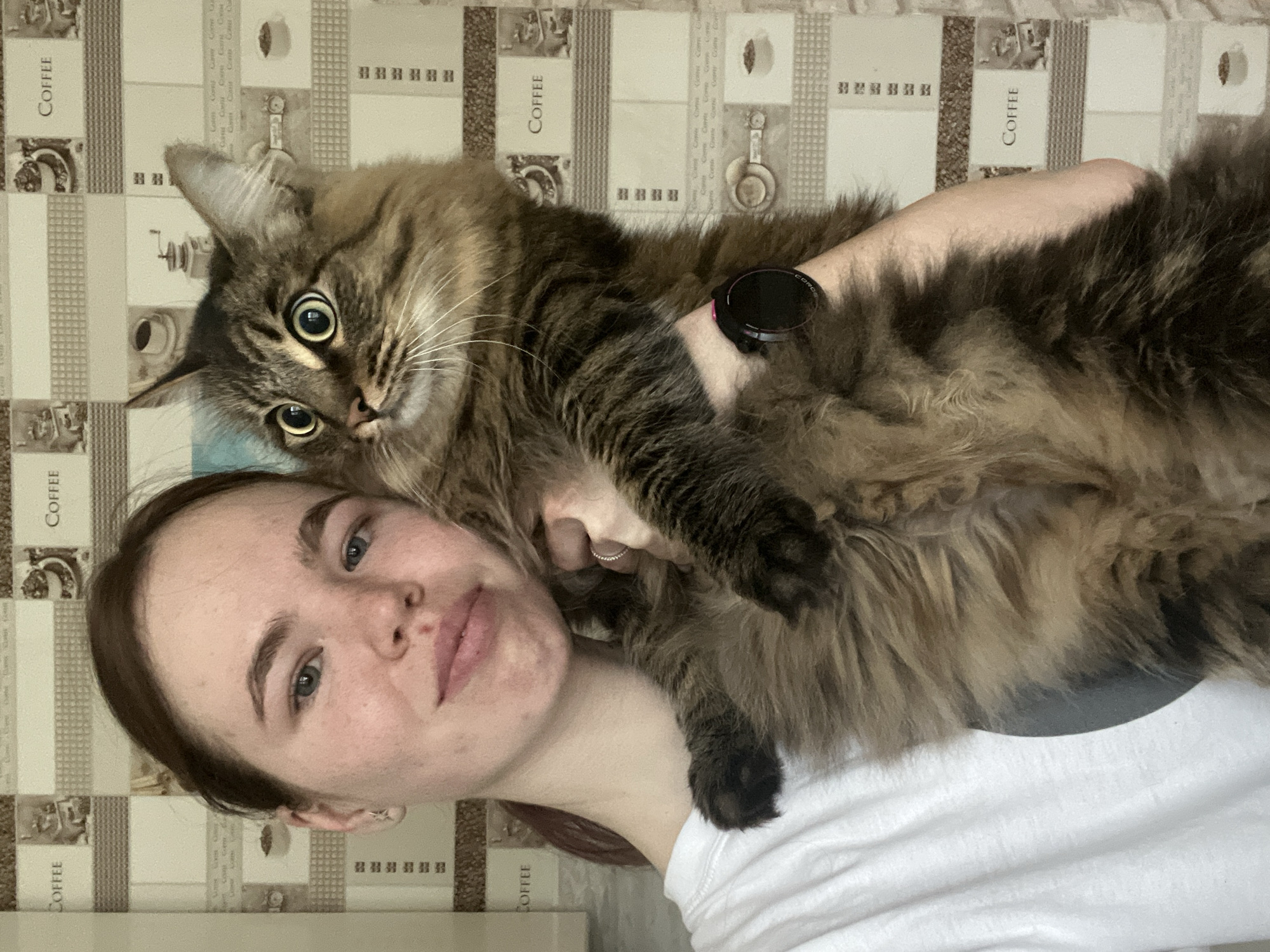 The image displays a young adult female holding a long-haired tabby cat indoors. The female, characterized by light brown hair pulled back and blue eyes, wears a white t-shirt and looks towards the camera with a slight smile. Her left hand, which has a ring on the ring finger, supports the cat, while a black Coros brand smartwatch with a pink accent is visible on her right wrist. The cat has brown and black striped fur, large green-yellow eyes with dilated pupils, and prominent white whiskers; its gaze is directed slightly to its left with an alert expression. The background is an interior wall covered with light-toned wallpaper featuring a repeating pattern of various coffee-related illustrations and the word "COFFEE." The specific location within the city of Chaikovsky, Russia, is not discernible from the visual evidence.