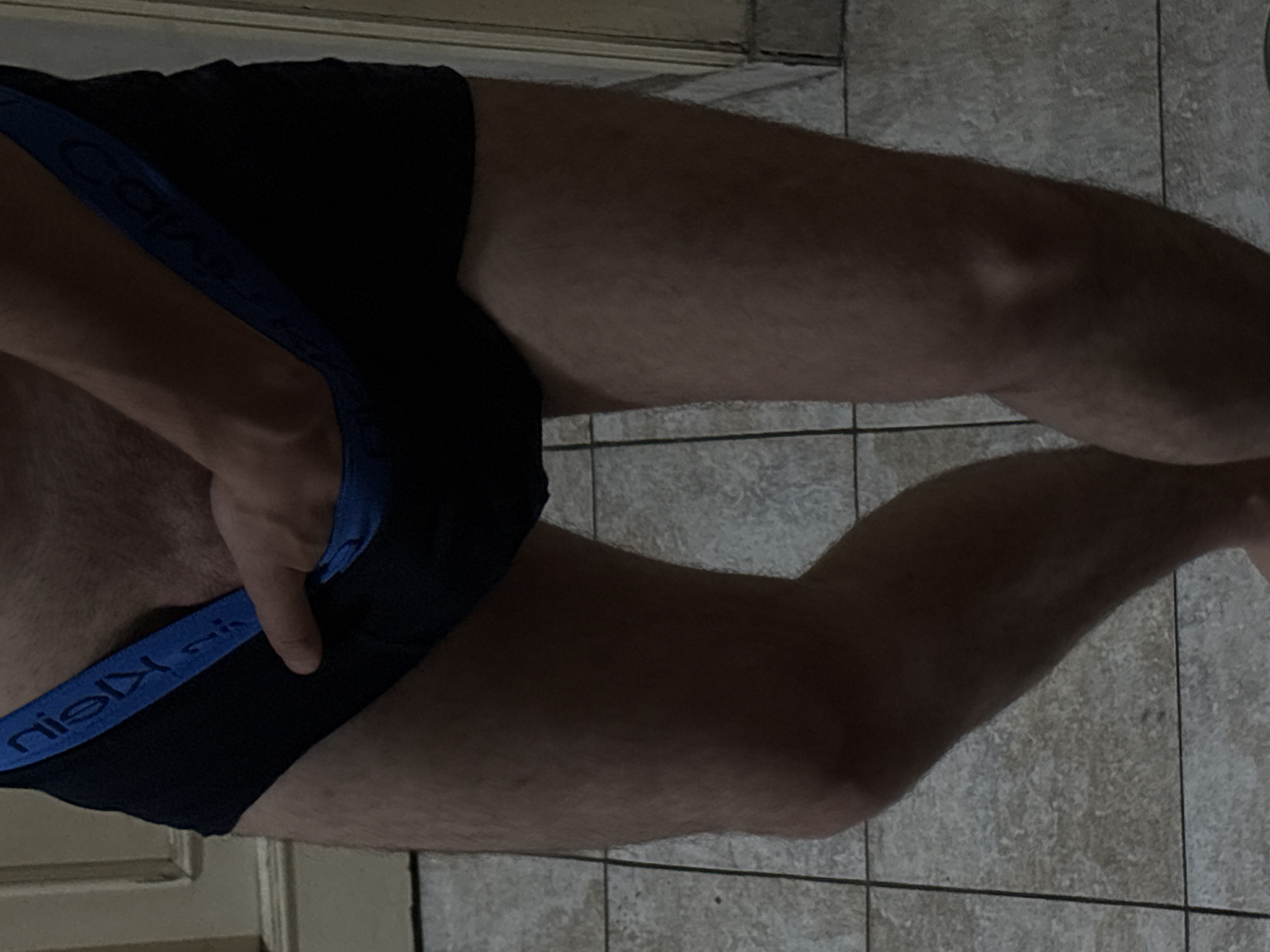 A close-up, indoor shot focuses on the lower torso and legs of a person wearing black Calvin Klein briefs with a blue waistband. A hand is visible, holding the briefs. The background shows a tiled floor and a door. The location is not discernible from the provided image.