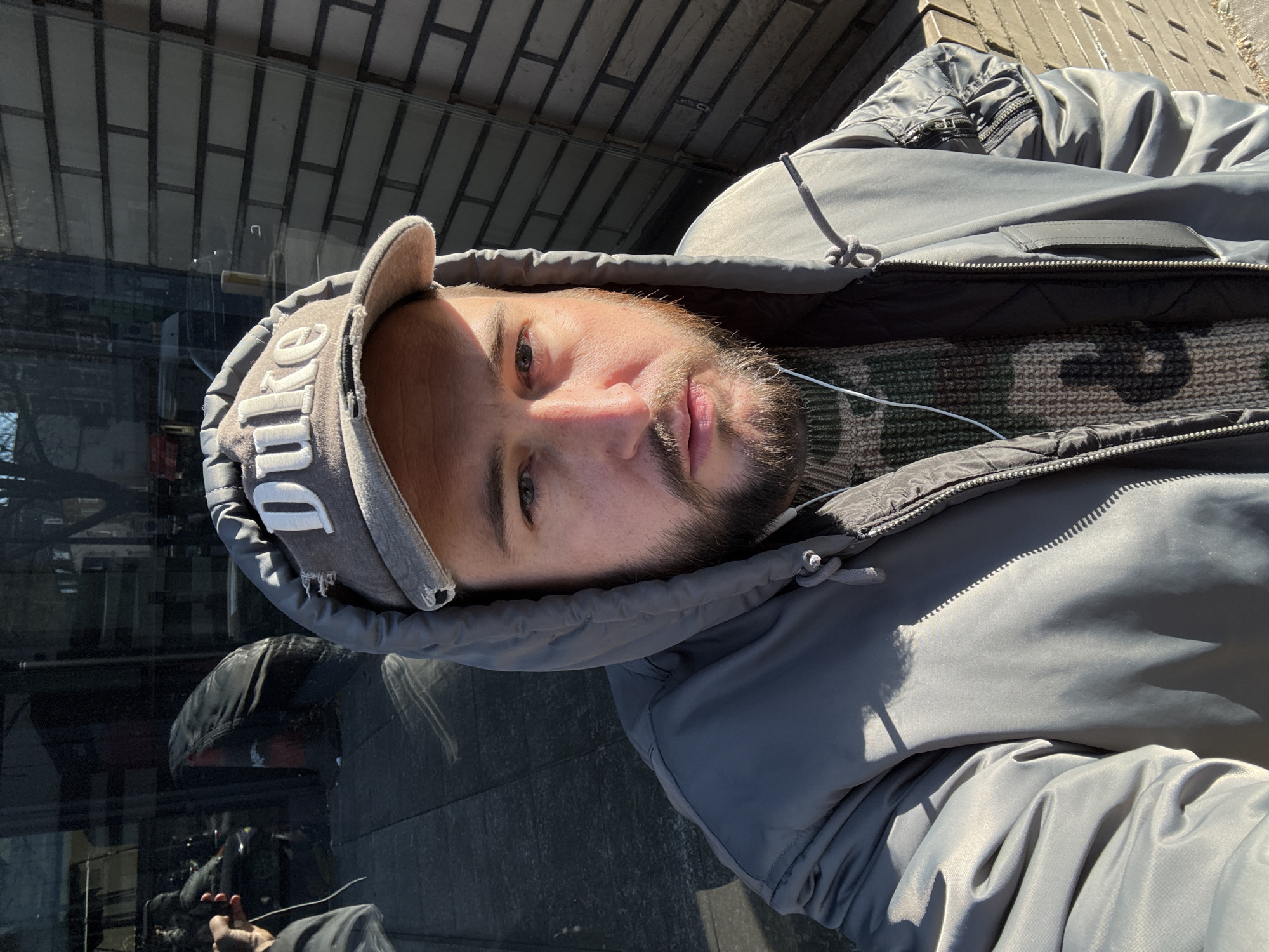 A man is seen from a top-down perspective, wearing a grey hooded jacket and a baseball cap with "DUKE" embroidered on it. He is possibly taking a selfie. His face is visible, showing a short beard and light stubble. He's wearing earphones. Under the jacket, a knitted, patterned sweater can be seen. The background features a reflective glass surface and a brick wall. Considering the 'DUKE' cap, the location may not be in London, United Kingdom. It is more likely in Durham, North Carolina (USA) where Duke University is located. Further evidence to verify the location needs a broader view.