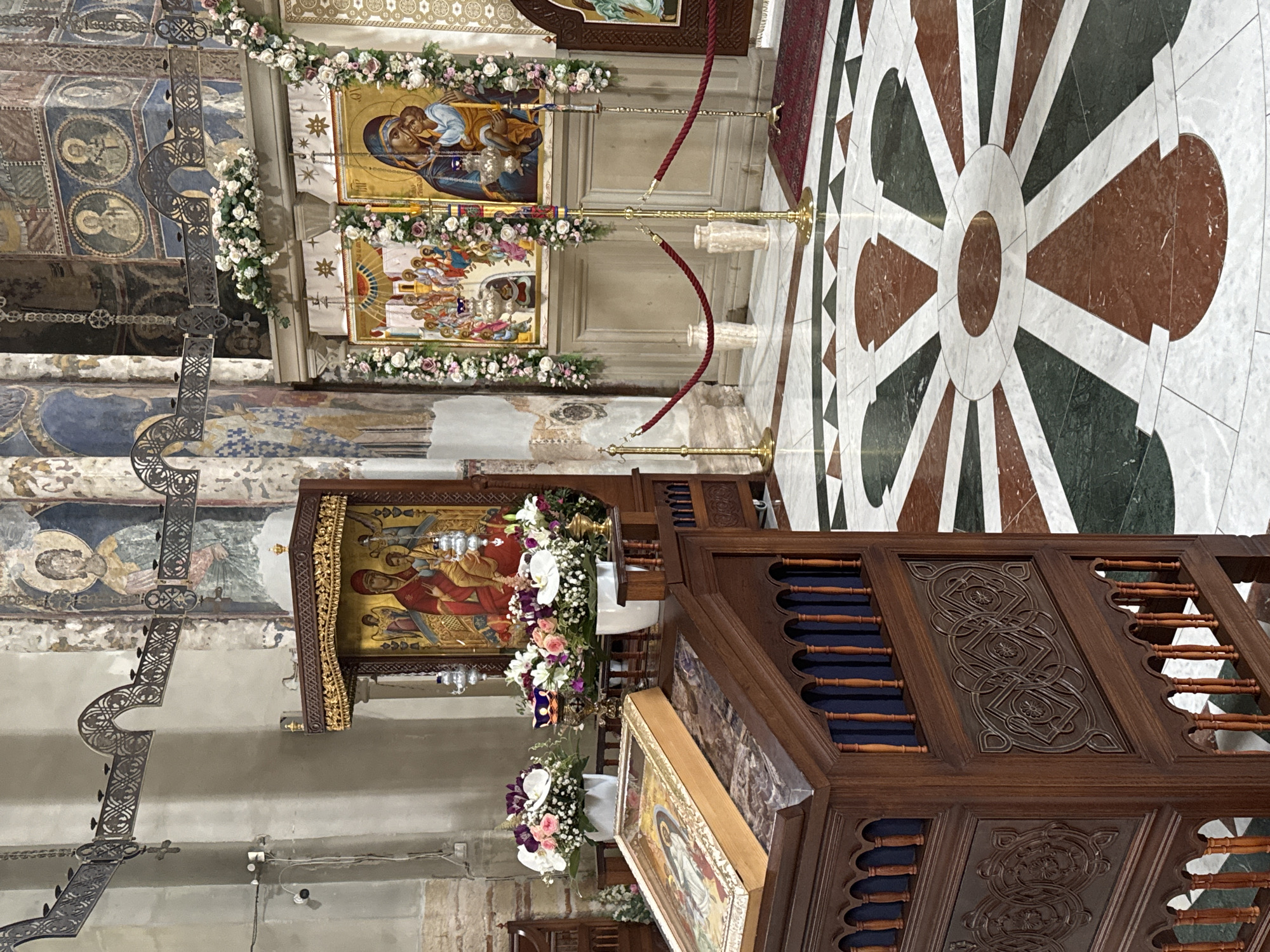 This appears to be the interior of the Manasija Monastery located in Despotovac Municipality, Serbia. The image shows various religious icons adorned with flowers, a wooden pulpit with intricate carvings, and a decorative marble floor. The walls and ceilings exhibit Byzantine-style frescoes. There are no people visible in the frame.