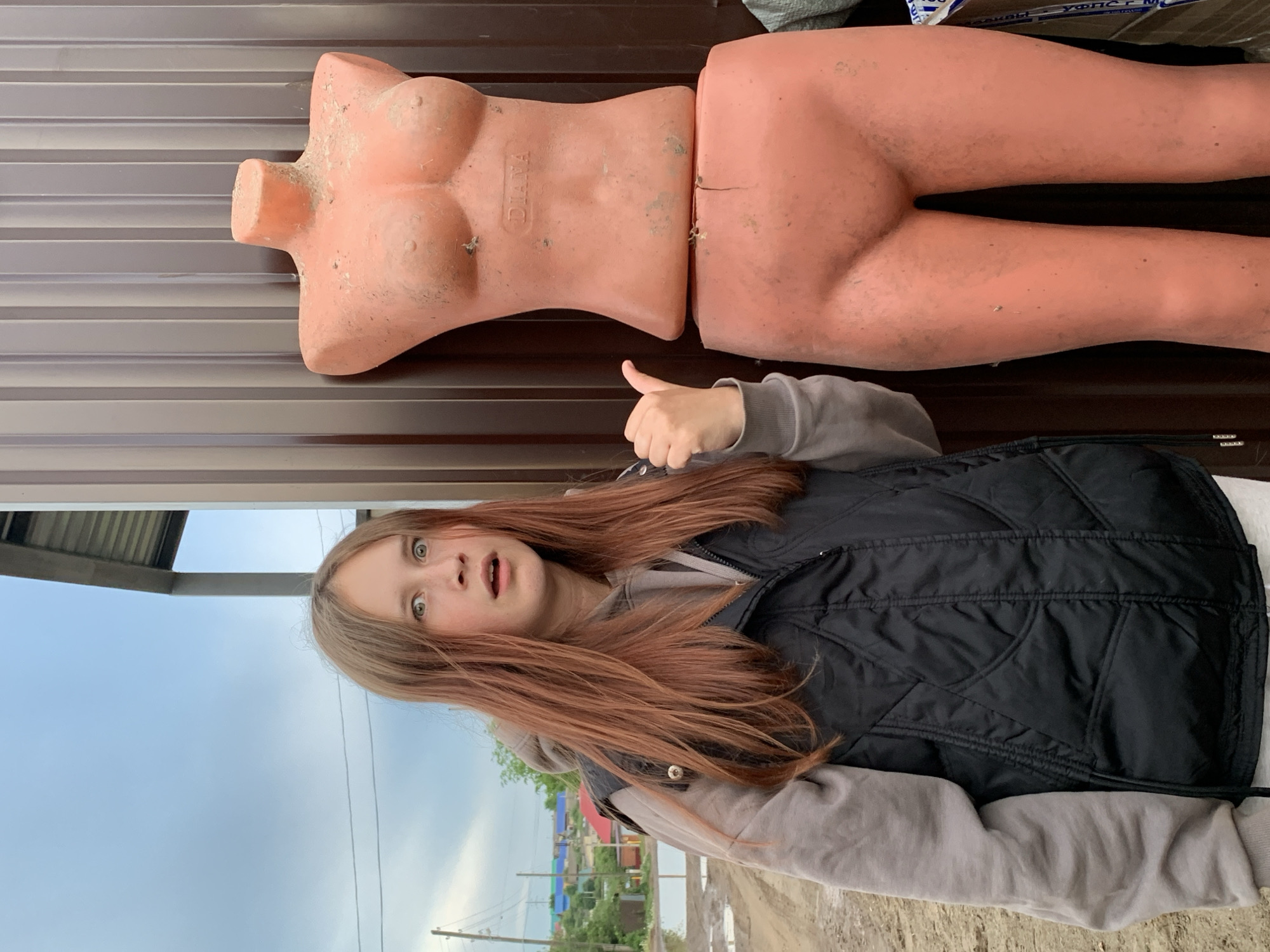 A young girl stands outside with a surprised expression, giving a thumbs-up gesture. She is wearing a grey hooded sweatshirt and a black vest. Behind her is a two-part, slightly dirty, flesh-colored mannequin torso and leg section. The mannequin appears to be lying on a horizontal surface, possibly a fence. Beyond the girl and fence, there is a view of Фоки, Russia, with houses and utility poles visible under a cloudy sky.