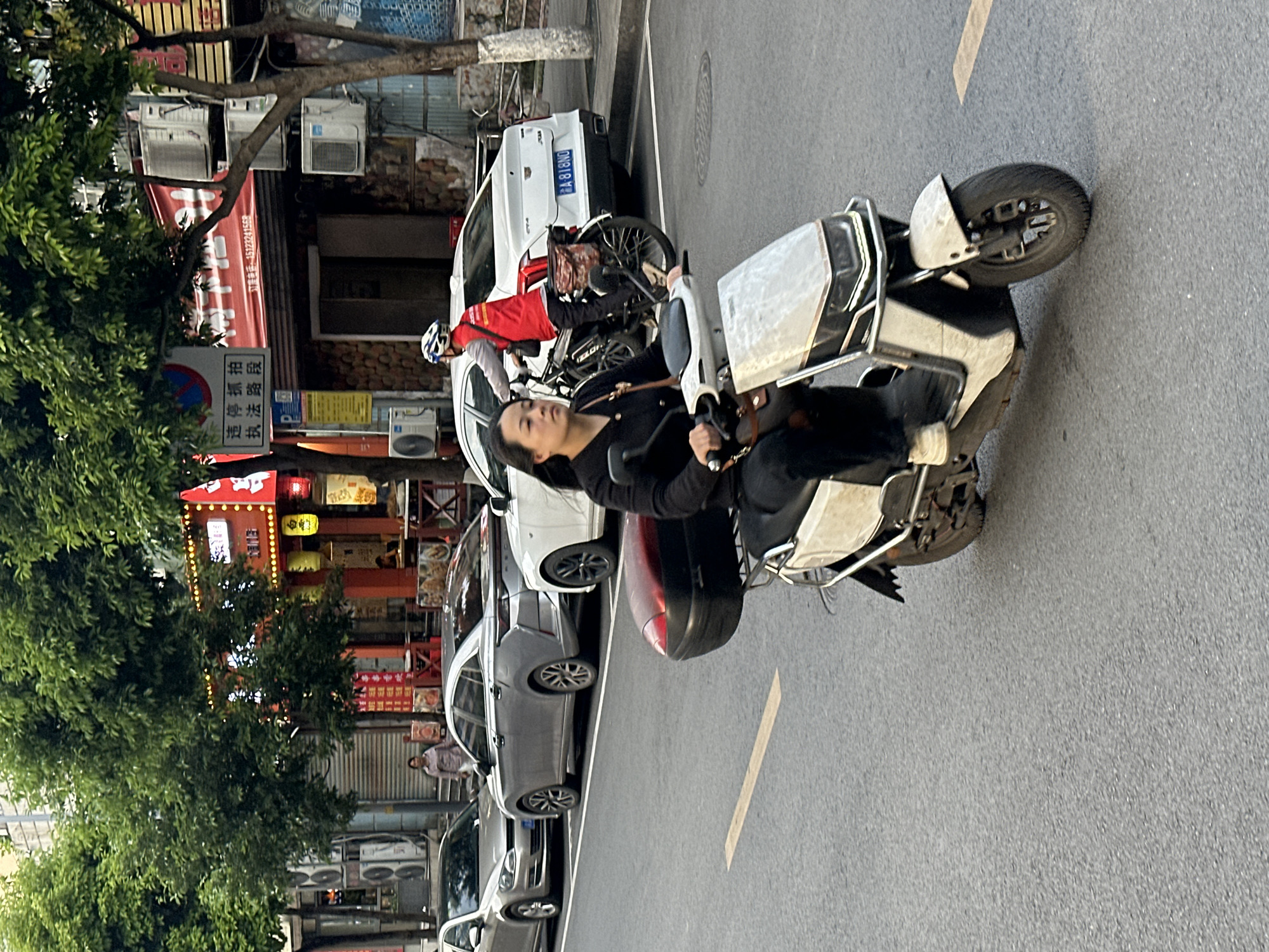 This image captures a bustling street scene in Jiangjin, China, likely during daylight hours with bright, possibly overcast, conditions. The main subject is a person, appearing to be a woman, with dark hair, wearing a black long-sleeved top and dark pants, seated on a white and silver electric scooter. She holds the handlebars and looks slightly to her right. The scooter itself has a front shield and a dark red component near the footrest area.
Behind the scooter and extending to the left, several cars are parked or moving slowly. Immediately behind the main subject is a white car, possibly an SUV, with a license plate partially visible as "A 818NO". A person in a red jacket and a white helmet is seen next to a bicycle, positioned behind the white car, suggesting a delivery rider. Further back, a grey sedan and another silver/grey car are visible.
The background features buildings lining the street, which appear to house shops or restaurants. Numerous outdoor air conditioning units are mounted on the walls of these buildings. Lush green foliage from trees partially obscures parts of the architecture. Red banners and signs with Chinese characters are prominent, and some decorative yellow and red lanterns hang from an establishment. The road is asphalt with a faded yellow line marking.
Visible text includes a phone number "13172241558" on a red banner, along with Chinese characters that translate to "Illegal photography, law enforcement section" or "Illegal parking enforcement section," indicating a regulated area. The overall scene depicts typical urban activity, with people commuting on scooters and bicycles alongside parked cars, set against a backdrop of local businesses.