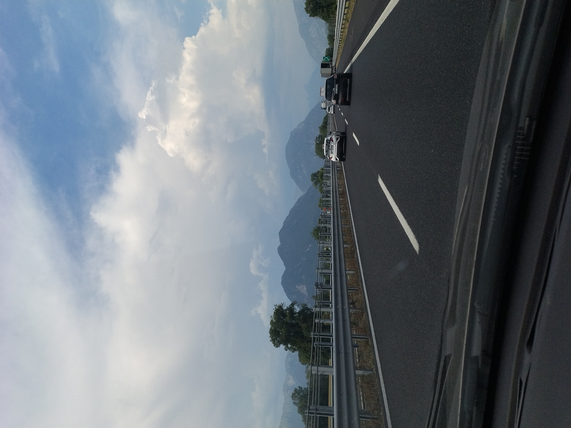 The image depicts a view from inside a vehicle traveling on a multi-lane highway. The highway surface is dark asphalt with white lane markings. A metal guardrail runs along the left side of the road. In the distance, a range of mountains forms the backdrop, with some appearing rocky and others covered in vegetation. The sky is a mix of blue sky and large, fluffy white clouds. Several vehicles are visible on the highway, including cars and a truck. The foreground shows the interior of the vehicle, including the windshield and dashboard. The scene suggests a journey through a scenic, mountainous region. The location is identified as being in the city of Buja, Italy, which is known for its proximity to mountainous landscapes and natural beauty, consistent with the visual elements in the image.