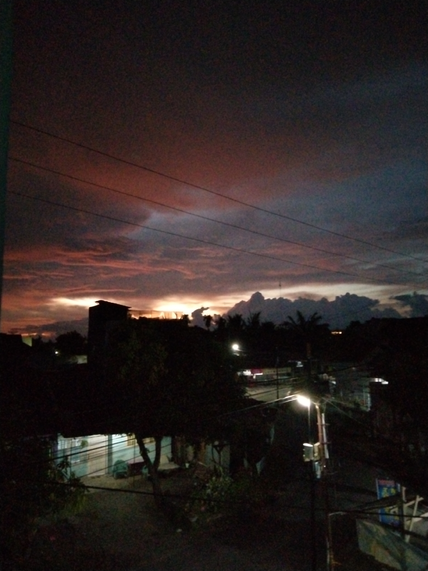 The image shows a night scene in Bireuenmeunasahdayah, Indonesia. The sky exhibits a gradient of color from a deep, dark red-tinged mauve to an inky black, indicating either a late sunset or early sunrise. Cloud formations are visible, some illuminated by the fading light. The horizon line is populated by silhouettes of buildings and trees. In the foreground is a residential area with indistinct shapes that suggest houses and dense foliage. A street lamp illuminates a portion of the scene, revealing power lines. The overall scene is dimly lit, creating a sense of depth and shadow.