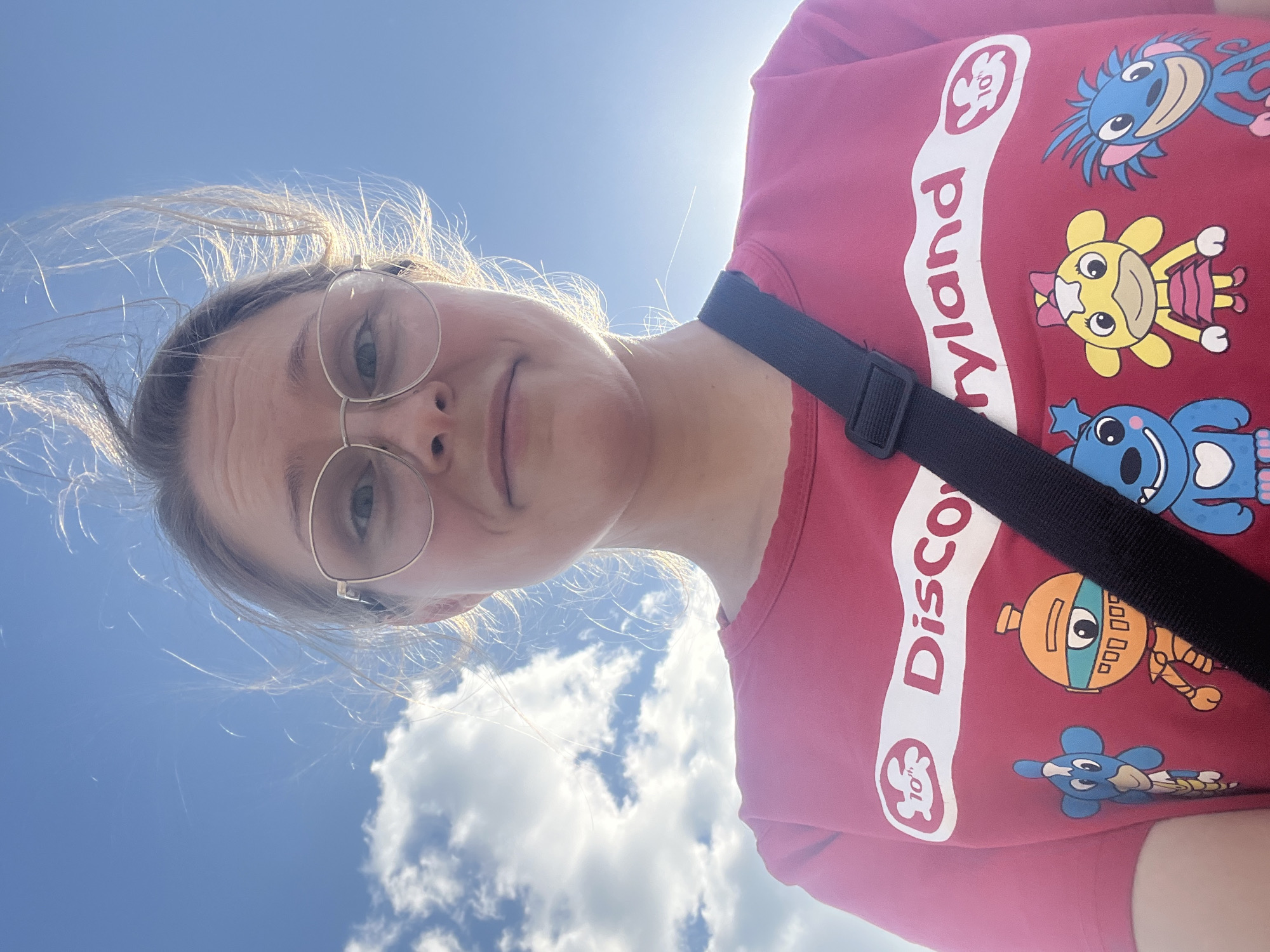 In Amel, Belgium, a young woman with blonde hair and glasses takes a selfie outdoors under a bright blue sky with scattered clouds. She is wearing a pink t-shirt with the word "Discoveryland" printed on it in white letters, along with cartoon characters and a "10th" anniversary logo. The woman's hair is blown by the wind. A black strap of a bag is visible across her t-shirt. The photo is taken from a low angle, looking up at the woman and the sky.