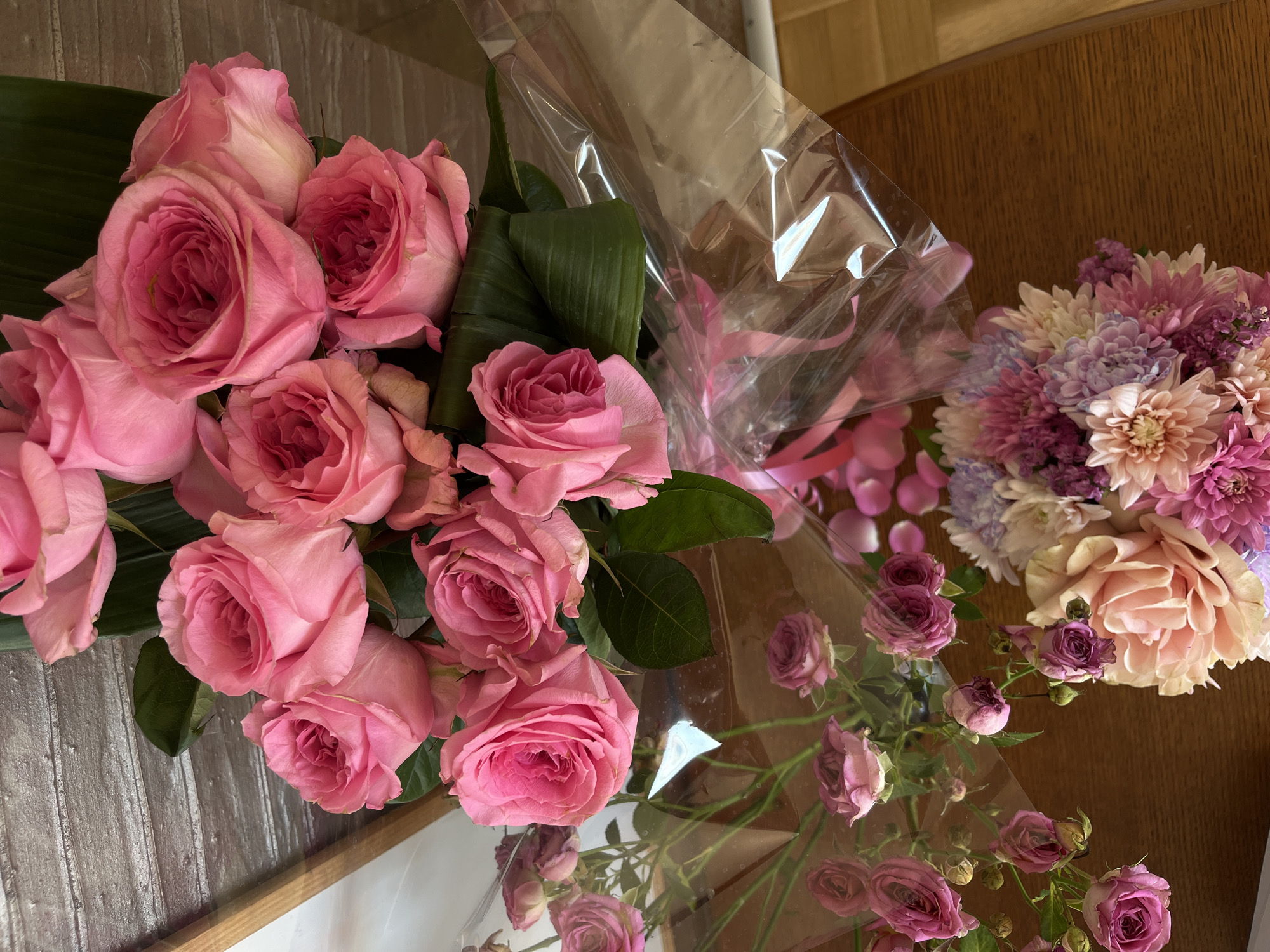 An overhead, close-up shot captures multiple bouquets of flowers arranged on a wooden table, likely indoors in Bečići, Montenegro. The prominent subject is a large, vibrant bouquet of fresh pink roses, wrapped in clear cellophane and positioned on the left side of the frame. These roses vary in shade from deep fuchsia to softer pinks, with several dark green leaves, possibly banana leaves, providing a contrasting backdrop.
To the right, a smaller, rounder bouquet features a mix of pink, purple, and cream-colored chrysanthemums or similar composite flowers, also partially encased in clear plastic. Scattered around this bouquet on the table are several loose pink flower petals. In the lower right corner, another arrangement consists of small, delicate spray roses, primarily in shades of deep pink and purple. Some of these smaller roses show signs of slight wilting, indicating they might be a little older. This bouquet is also wrapped in clear cellophane.
The wooden table surface, with its visible grain, serves as a warm, natural backdrop for the colorful floral display. The lighting is soft and even, suggesting a daytime setting. There is no visible text in the image. The collection of bouquets implies a festive occasion, a gift, or a decorative arrangement.