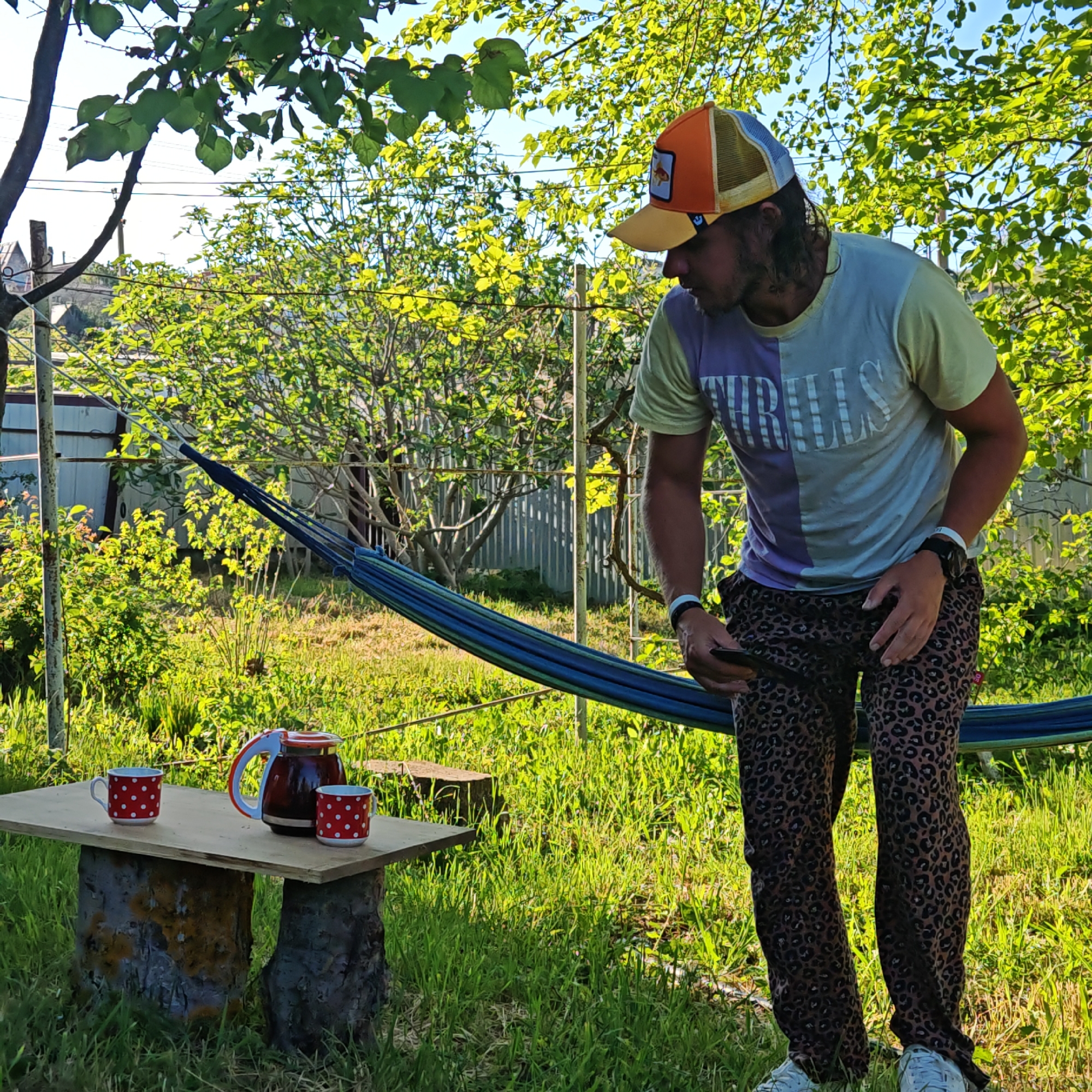 A man in an orange and yellow hat, a t-shirt, and leopard-print pants stands in a grassy yard. He is looking down and to his right, and is holding a phone in his left hand. Behind him is a blue and green hammock strung between two metal poles. In front of him, to the left, is a small wooden table resting on two tree stumps. On the table are two red and white polka-dotted mugs and a dark brown carafe with a red lid. The yard is filled with lush green grass and various trees and bushes. A white fence and a metal storage shed are visible in the background. Overhead, tree branches with green leaves provide partial shade. The location appears to be a backyard or garden setting in a residential area.