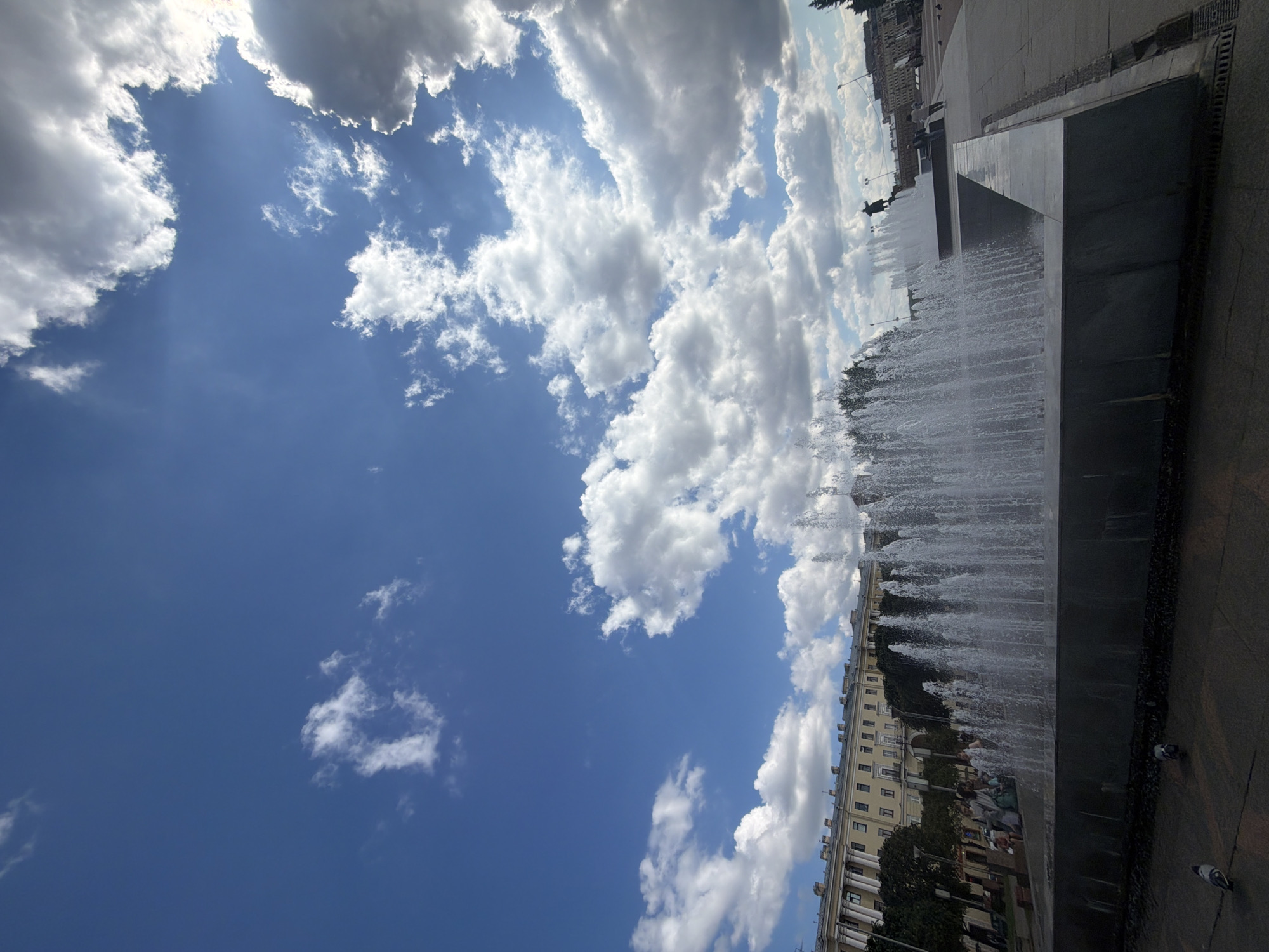 In Saint Petersburg, Russia, the image captures a wide-angle view of the city's vibrant public space. A large fountain dominates the foreground, its water jets creating a dynamic visual element against the backdrop of a majestic building and a bright blue sky dotted with fluffy white clouds. In the distance, a statue of a historical figure stands tall, silhouetted against the sky, drawing the viewer's attention to another point of interest within the urban landscape. The scene is further enlivened by the presence of several people, some walking and others seated, adding a sense of everyday life to the grandeur of the architecture and the natural beauty of the sky. The overall impression is one of a lively, sun-drenched day in a historically rich city.