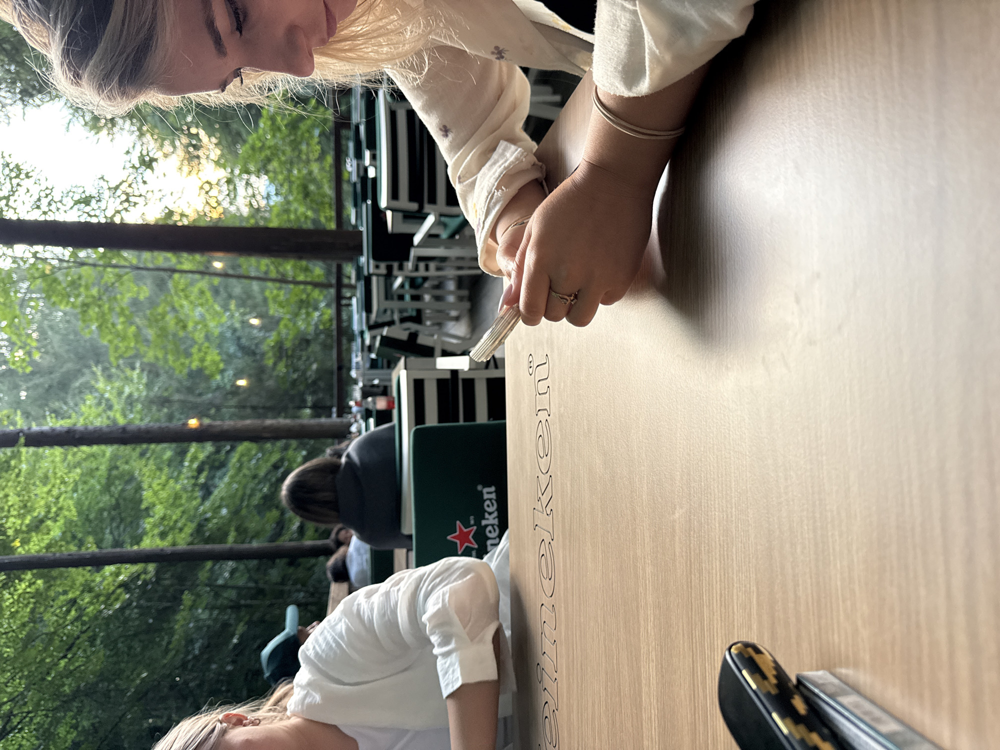 In Arefu, Romania, an outdoor dining area is depicted with a focus on two young women at a table. The woman on the right, with blonde hair and light-colored clothing, is looking down at her phone, her hands resting on a wooden table with "Heineken" branding. She wears rings on her fingers and a bracelet on her wrist. The woman on the left, partially visible, also wears light-colored clothing. In the background, other patrons are seated at tables, and the area is surrounded by tall trees, suggesting a natural, possibly forested, setting. The lighting indicates it is likely late afternoon or early evening, with sunlight filtering through the trees.