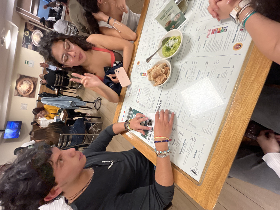A group gathers around a restaurant table covered in a menu. A young woman with glasses flashes a peace sign while another leans in. On the table, there are dishes of guacamole and chicharrones. A young man at the table holds a phone, possibly taking a picture. Other patrons sit at tables in the background, and a TV hangs on the wall.