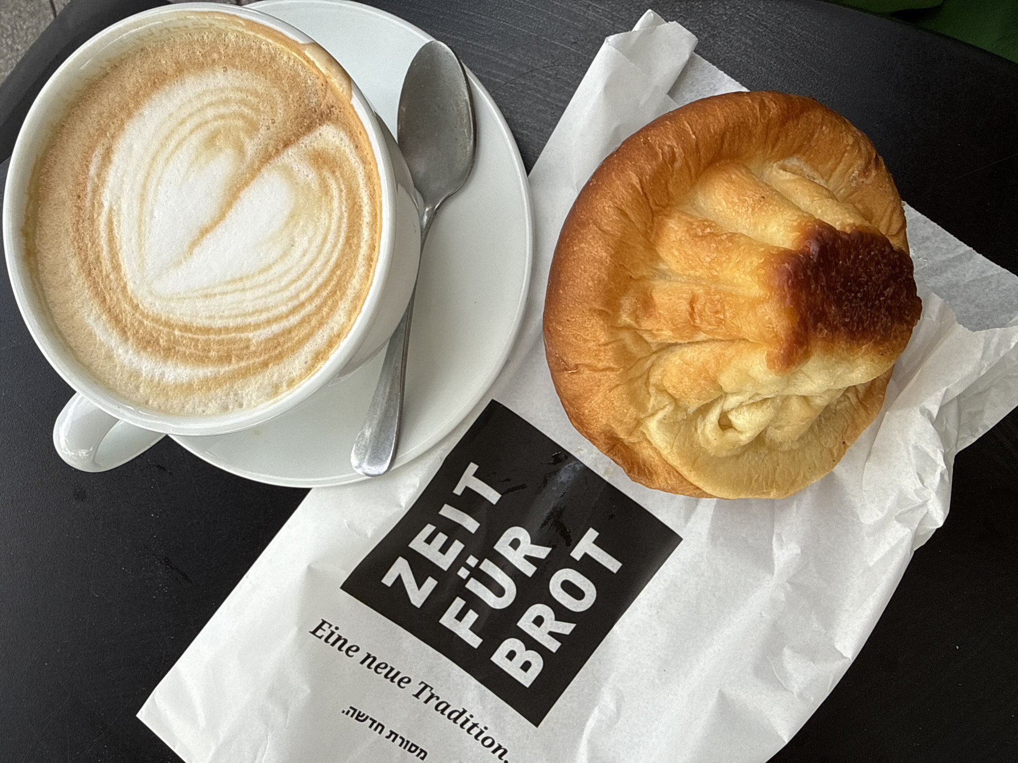 A high-angle, medium shot captures a cup of cappuccino, a pastry, and a bag from the bakery "Zeit für Brot." The cappuccino, decorated with latte art, sits on a white saucer next to a silver spoon. The pastry, golden brown with a dark, glazed top, rests on a sheet of white paper. Adjacent to the pastry is a partially visible white paper bag, printed with a black square diagonally displaying "ZEIT FÜR BROT" in bold, capital letters. Below the square, "Eine neue Tradition." is printed, along with Hebrew text. The composition sits on a dark surface. The image hints at the location being Tel-Aviv, Israel, given the Zeit für Brot branding which has a branch in the city.