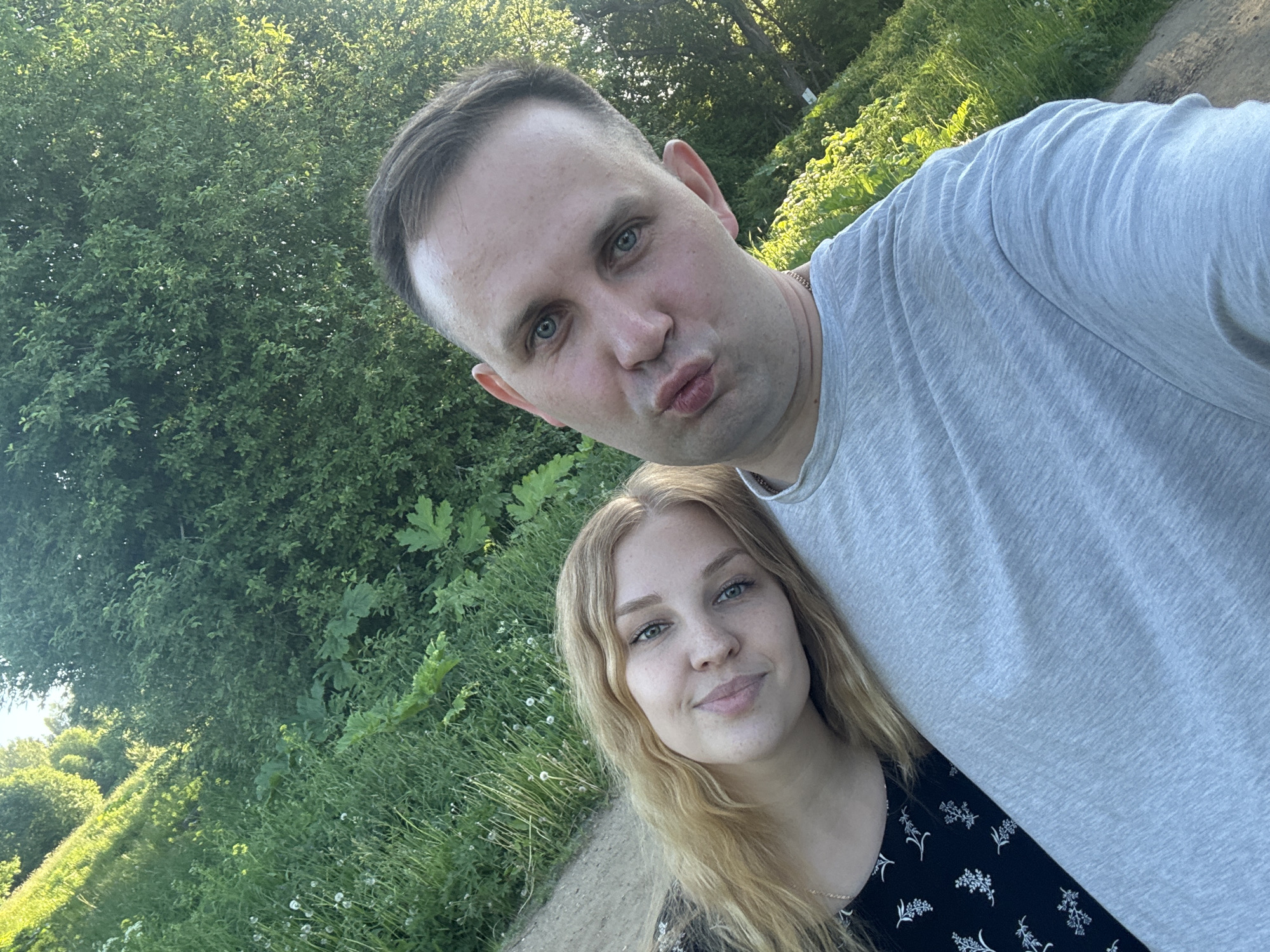 Two individuals, a male and a female, are captured in a selfie in an outdoor environment in Lepilovo, Russia. The camera is held by the male, positioned slightly above and angled downwards.
The male, on the right, has short dark hair with lighter sides and blue eyes. He wears a light grey short-sleeved t-shirt and a thin chain around his neck. His lips are puckered in a "duck face" expression as he looks directly at the camera.
The female, on the left, has long wavy blonde hair, blue-green eyes, and light freckles. She wears a black garment with a white floral pattern. Her head is tilted and rests on the male's left shoulder, and she exhibits a subtle smile while looking towards the camera.
The background features dense green vegetation, including tall grass with visible dandelion seed heads, various bushes, and trees, all illuminated by bright natural sunlight. A narrow dirt or gravel path runs along the left side of the frame behind the female.