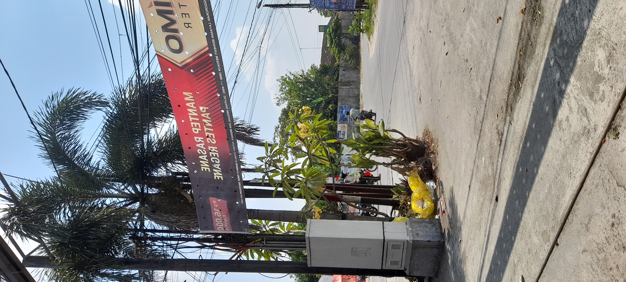 In the city of Kalisari, Indonesia, this image captures a street scene on a sunny day. A large red and gold banner hangs overhead, displaying text in what appears to be Indonesian, with some letters reversed. The banner advertises "MANTAP RAGANE" and "MANTAP GULING," with the text "OMINO" and "TER" also visible on the right side, possibly indicating a business or event.
Beneath the banner, palm trees with their fronds reaching towards the blue sky are prominent. A white electrical or utility box stands on a concrete base to the left of the frame, with yellow plastic bags and debris scattered at its base.
The street itself is paved with asphalt, and a motorcycle is parked on the left side of the road. A person in a yellow vest, likely a traffic controller or worker, stands near the motorcycle. Further down the road, a person is riding a motorcycle away from the camera. The environment includes some greenery, buildings, and overhead electrical wires. The overall impression is of a typical day in a commercial or residential area of Kalisari.