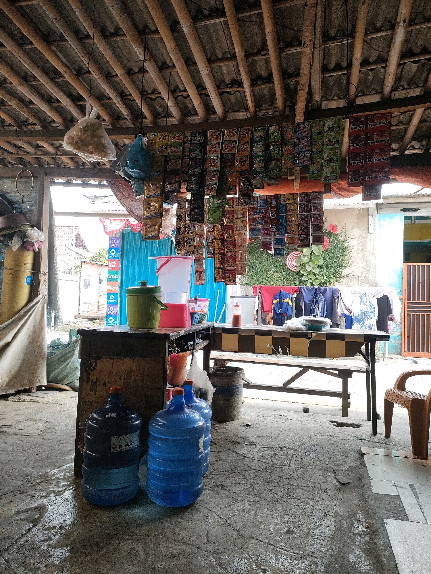 The image depicts an outdoor street-side stall in Karangasih, Indonesia. The stall is covered by a thatched roof supported by bamboo. Various goods are displayed, including sachets of food products hanging from the ceiling, a stack of blue water jugs in the foreground, a wooden counter with items on top, and laundry hanging on a clothesline behind the counter. The ground is concrete and appears wet in some areas. A plastic chair is visible to the right. The overall scene suggests a small, informal retail environment.