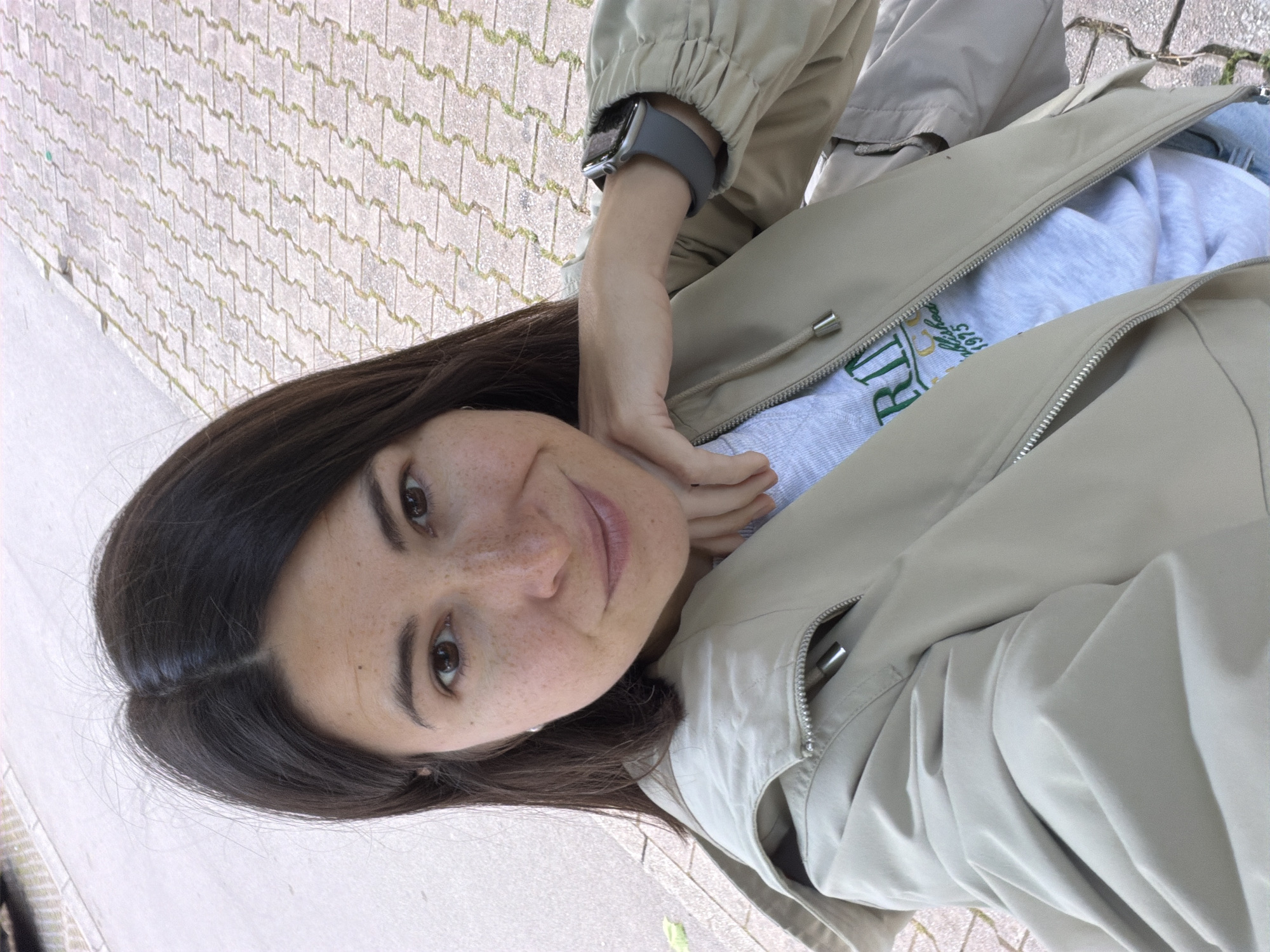 A portrait-style outdoor photograph captures a young woman from the chest up, looking directly at the camera with a gentle, relaxed smile. She has long dark hair, brown eyes, and freckles across her nose and cheeks. Her left hand is casually resting near her chin.
She is wearing a light beige or khaki zip-up jacket, which is partially open, revealing a light grey sweatshirt underneath. A grey smartwatch with a square face is visible on her left wrist. Partially visible green text on her sweatshirt reads "D 1975" and what appears to be "RI" or "RLD".
The background features a paved ground, likely a sidewalk or pathway, composed of interlocking light-colored pavers with some grass or moss growing in the cracks. The lighting is bright and even, indicating it is daytime. The photo was taken in Budva, Montenegro.