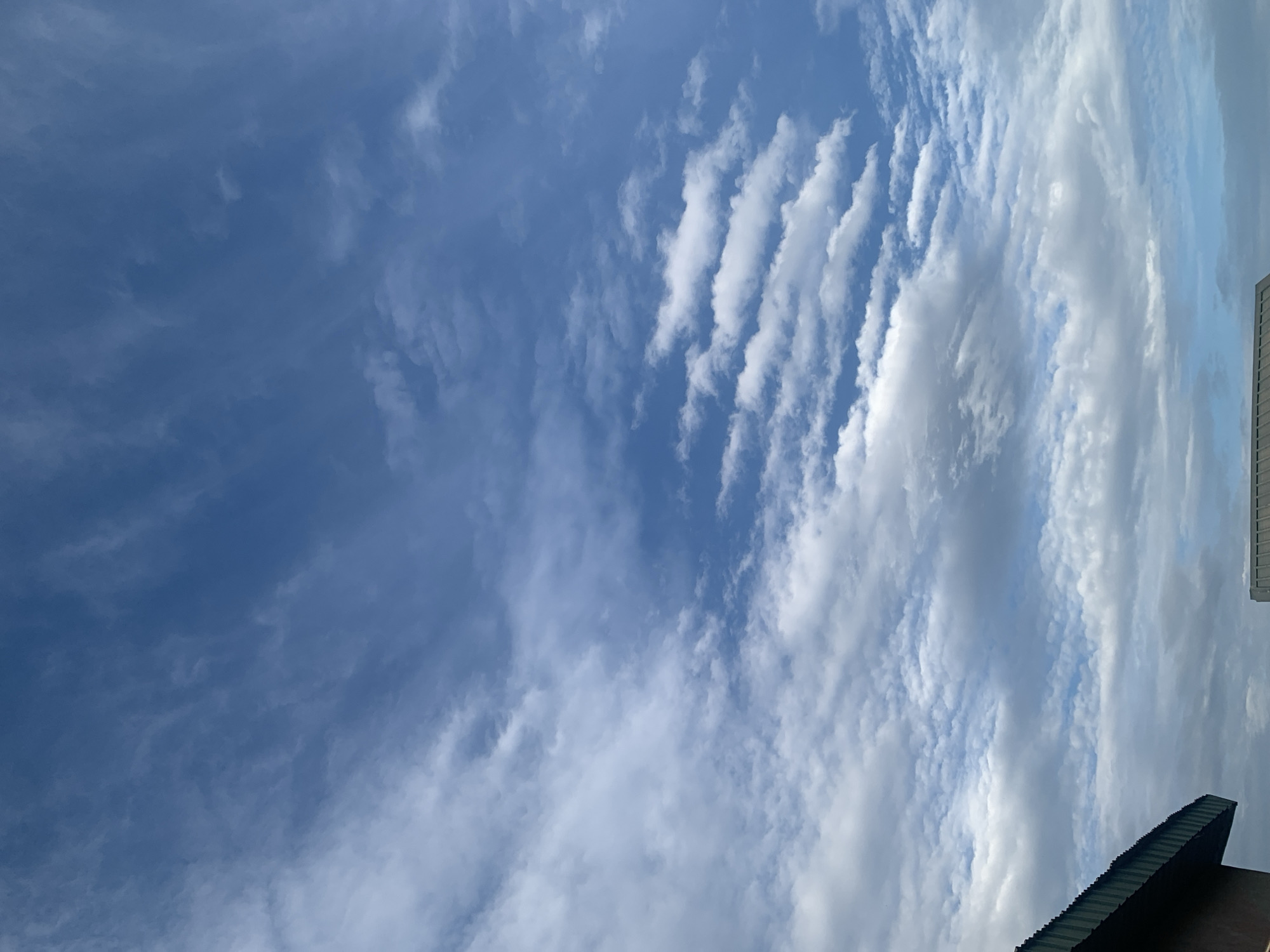 The image captures a sky filled with a dynamic mix of clouds over Sabon Tasha, Nigeria. Wispy cirrus clouds stretch across the upper portion of the frame, interspersed with scattered cumulus clouds that form distinct rows and patterns in the middle ground. The lower part of the image shows a sliver of a building with a dark green, corrugated metal roof in the bottom left corner, and a hint of another structure with a similar roofline in the bottom right. The overall scene is dominated by the vast expanse of the sky, with a clear blue tone visible through the cloud formations.
