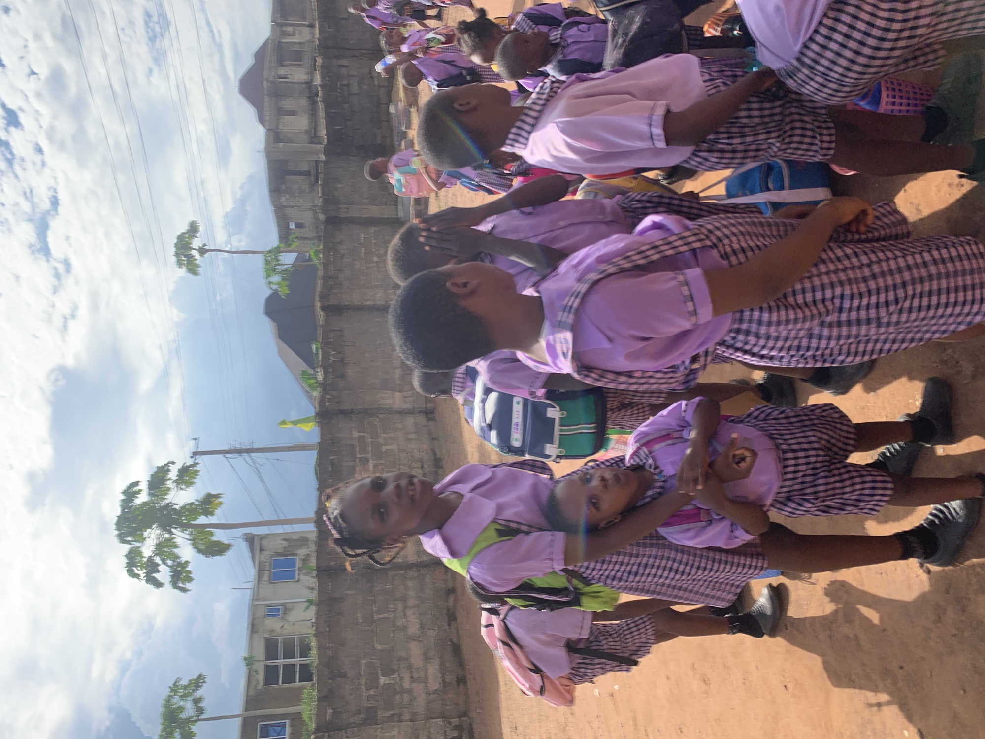 In Ukwu-Abwa, Nigeria, a group of schoolgirls in checkered dresses with purple tops sit outside on a sunny day. A brick building and trees can be seen in the background. Some girls are interacting with each other.