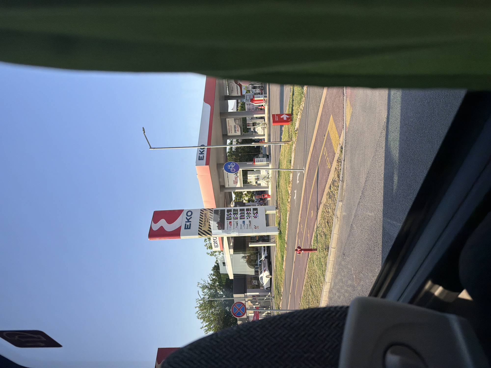 The image depicts a view of an EKO gas station, likely located in Plovdiv, Bulgaria, as suggested by the Bulgarian text on the price board. The gas station features a prominent EKO logo and red and white branding on its canopy. Several fuel pumps are visible, along with cars parked at the station. A price board displays current fuel prices, including Unlead 95, Diesel, and LPG, in Bulgarian currency. Road markings, including a "STOP" sign and a bike lane symbol, are present on the road in front of the station. A fire hydrant is also visible near the road. The scene is bathed in natural daylight, with a clear blue sky overhead, suggesting it is daytime. The photograph is taken from inside a vehicle, with parts of the vehicle's interior, such as a seat and a green curtain, obscuring portions of the view.