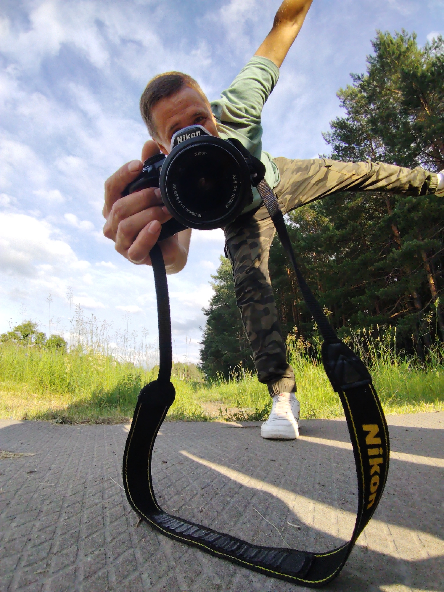 The image shows a person holding a Nikon camera at arm's length, as if taking a selfie or recording a video. The person is wearing a light green long-sleeved shirt and camouflage pants, with white sneakers. They are positioned outdoors in what appears to be a park or natural area, with green grass, trees, and a paved path. The sky is visible with clouds. The camera strap, with the prominent "Nikon" logo, is also visible. The low camera angle suggests the photo was taken from a crouching or very low standing position. The location is identified as Chaikovsky, Russia.