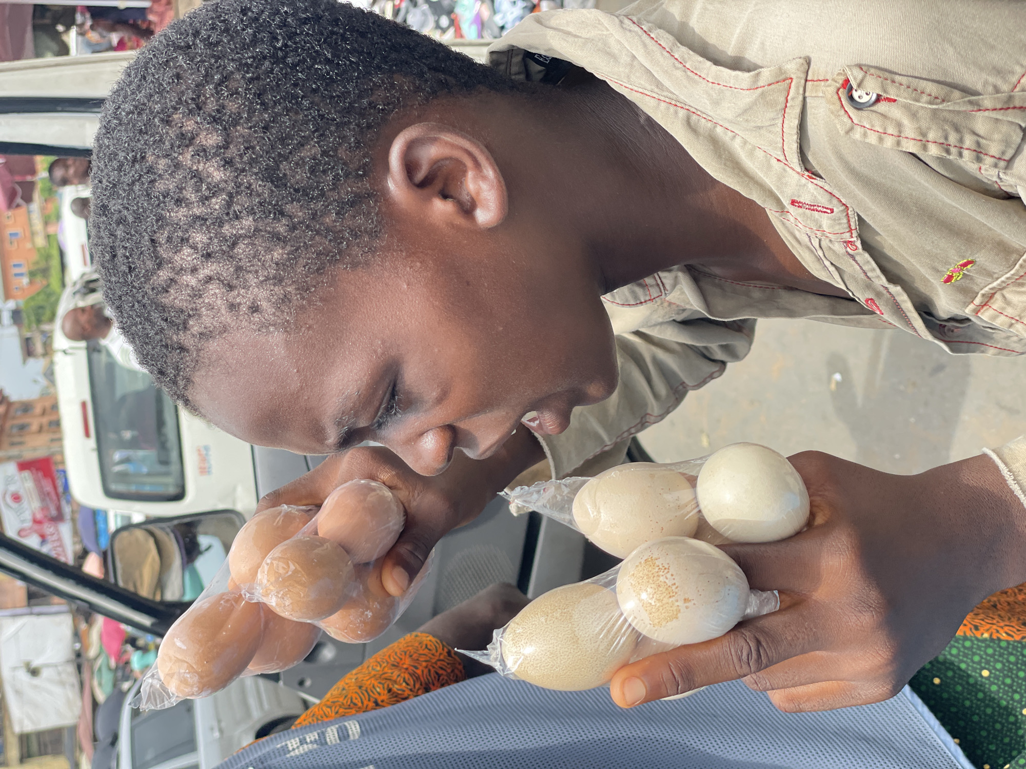 In an outdoor setting in Onitsha, Nigeria, a young Black male is holding two bags of eggs, one with brown eggs and the other with white eggs, both packaged in clear plastic bags. The man has a close-cropped haircut and wears a tan collared shirt with red stitching. He is looking down at the eggs in his hands. The background includes a white van, other people, and surrounding buildings. The man's skin is visible as brown.