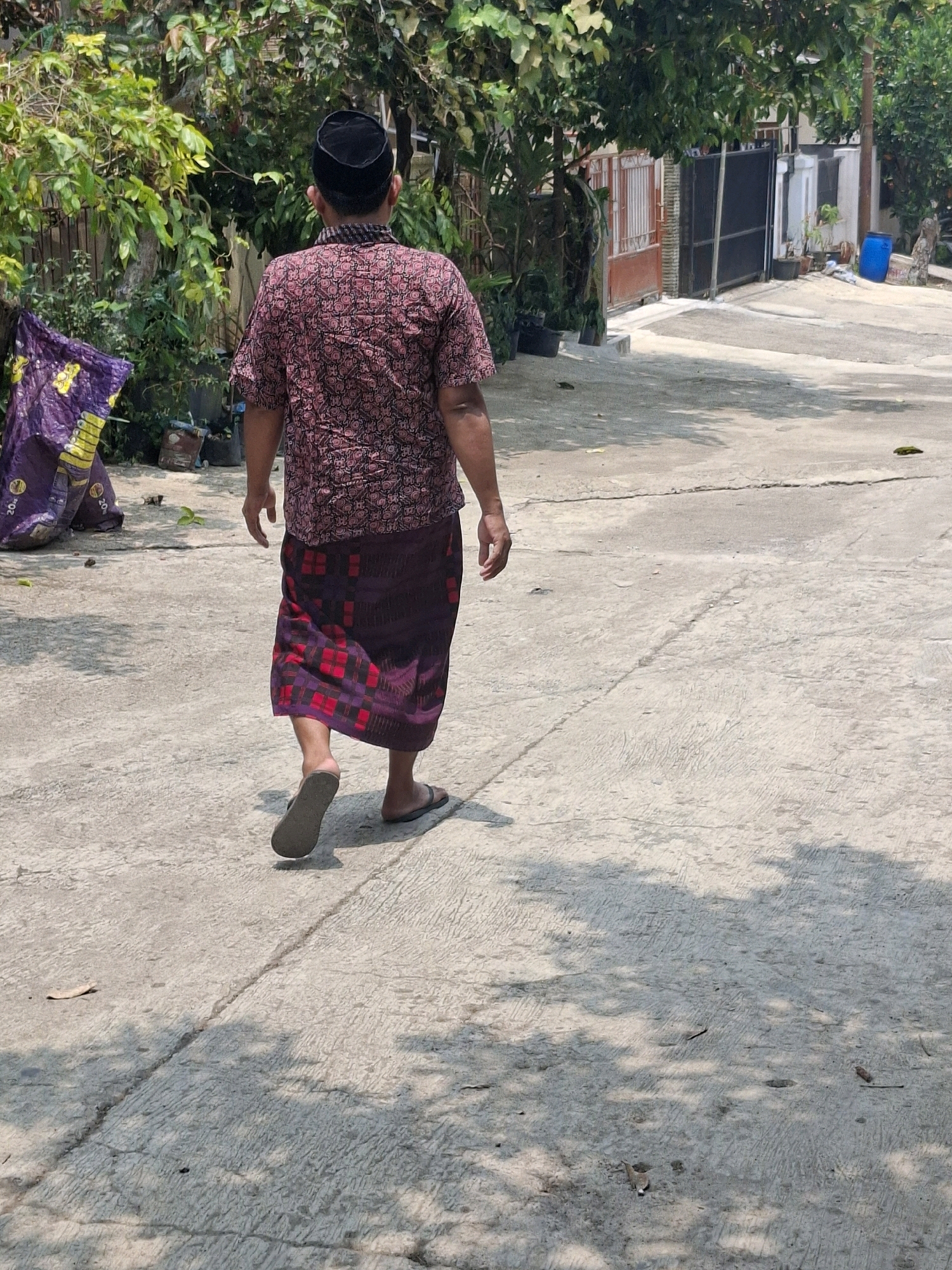 In Ciheulang Tonggoh, Indonesia, a man walks away from the camera down a concrete path. He wears a dark, brimless cap, a short-sleeved, patterned shirt in shades of red and pink, and a dark purple sarong with a red checkered pattern. His right foot is slightly raised, mid-stride, and he wears dark flip-flops. To the left of the path is a large, crinkled purple bag with yellow markings, placed near lush green foliage and trees. Further down the path, houses with closed gates are visible, including a red-striped gate and a black metal gate. A blue barrel and other debris are also present in the background. The ground is dappled with sunlight and shadows from overhead trees.