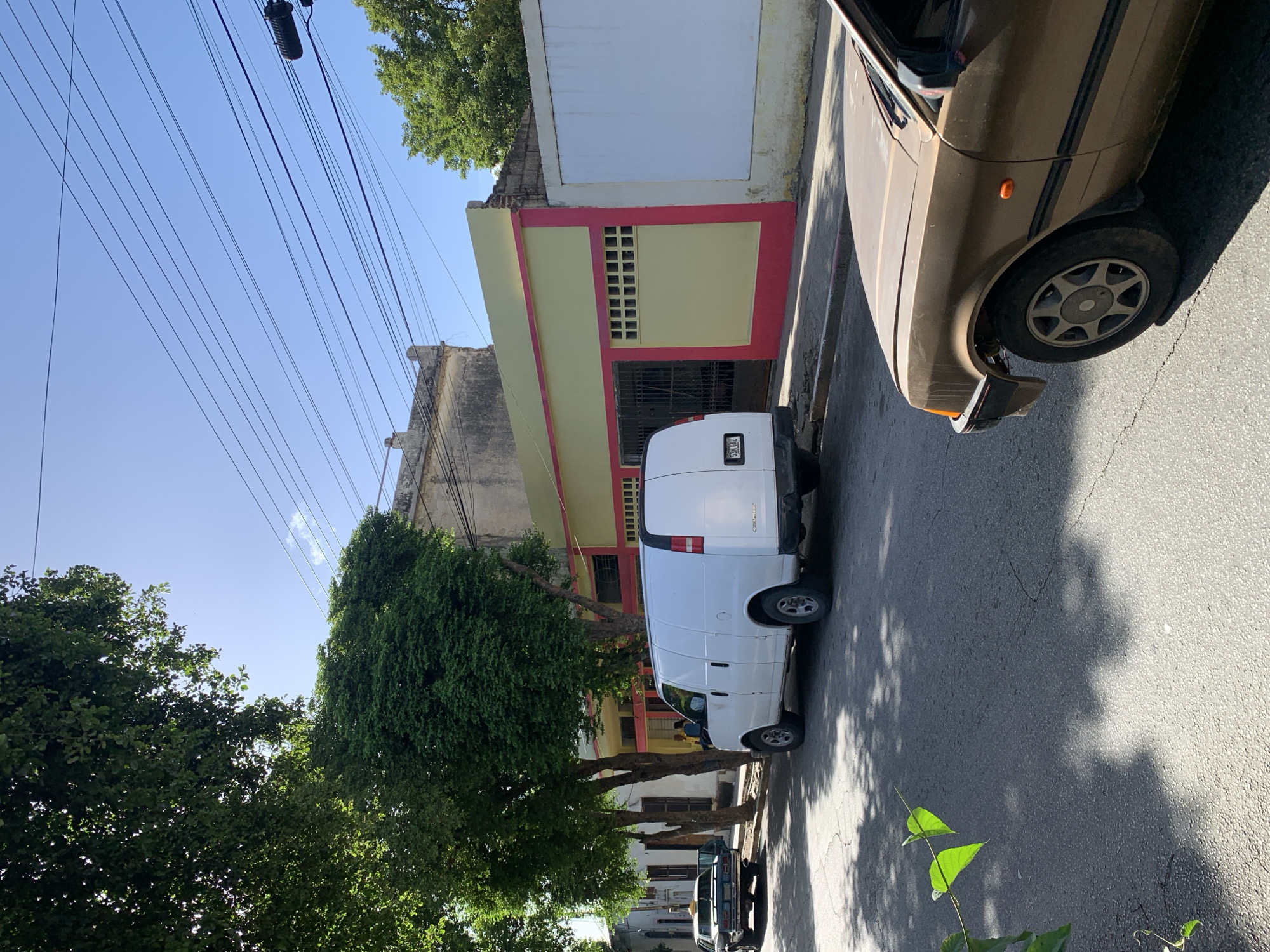This image captures a street scene in Santa Ana, Venezuela, under a bright, clear blue sky. In the foreground, the front section of a brown, older model car is visible, parked on a cracked asphalt road. To the left, a white cargo van is parked, its rear facing the camera. Beyond the van, a dark-colored car, possibly a taxi, is parked with its driver's side door ajar.
The street is lined with trees providing dappled shade on the road. Behind the trees, a building with a yellow facade trimmed in pink is visible. This building has metal gates and a section with a grid of small windows. To the right of this building is a white wall. Overhead, a complex network of electrical wires stretches across the sky, supported by poles. The overall impression is a quiet, residential street in a Venezuelan city on a sunny day.