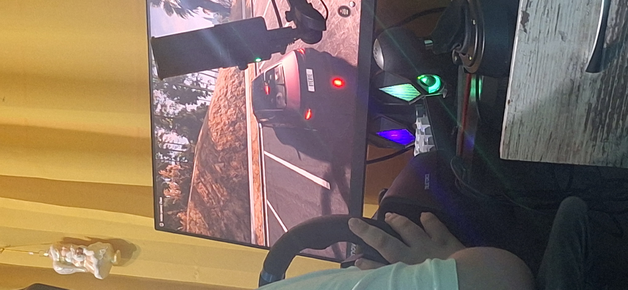 A person sits in front of a computer monitor, holding a gaming steering wheel. On the screen, a vehicle can be seen, indicating the person is likely playing a driving simulation video game. Based on the vehicle's license plate, it can be identified that the location in the game is Stara Zagora, Bulgaria. Next to the monitor on the right side there is a computer tower. On the wall, to the left of the monitor, there is a decorative object hanging. The person is wearing a light-colored shirt. A wooden surface is partially visible to the right of the computer tower. The room is lit by a warm, yellow light.